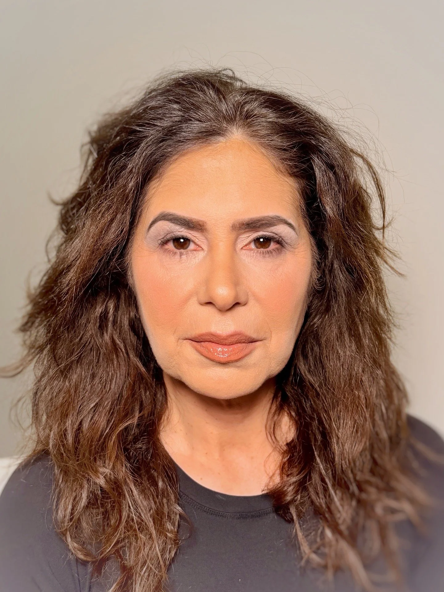 A woman with shoulder-length, wavy brown hair, wearing makeup, a black top, and neutral lipstick, looking directly at the camera against a plain background.