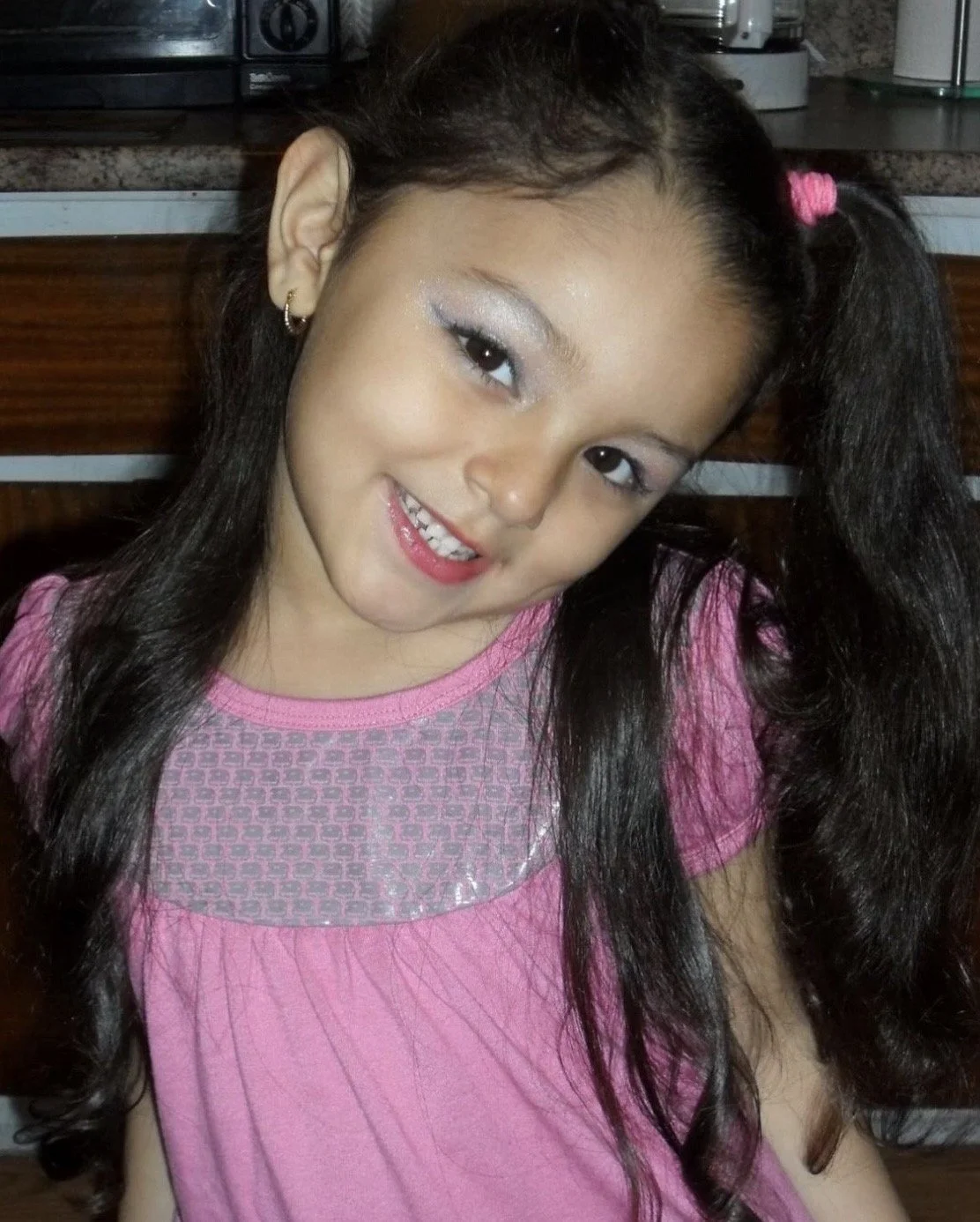 A young girl with long dark hair styled in pigtails, wearing a pink shirt, smiling at the camera, in a kitchen setting.
