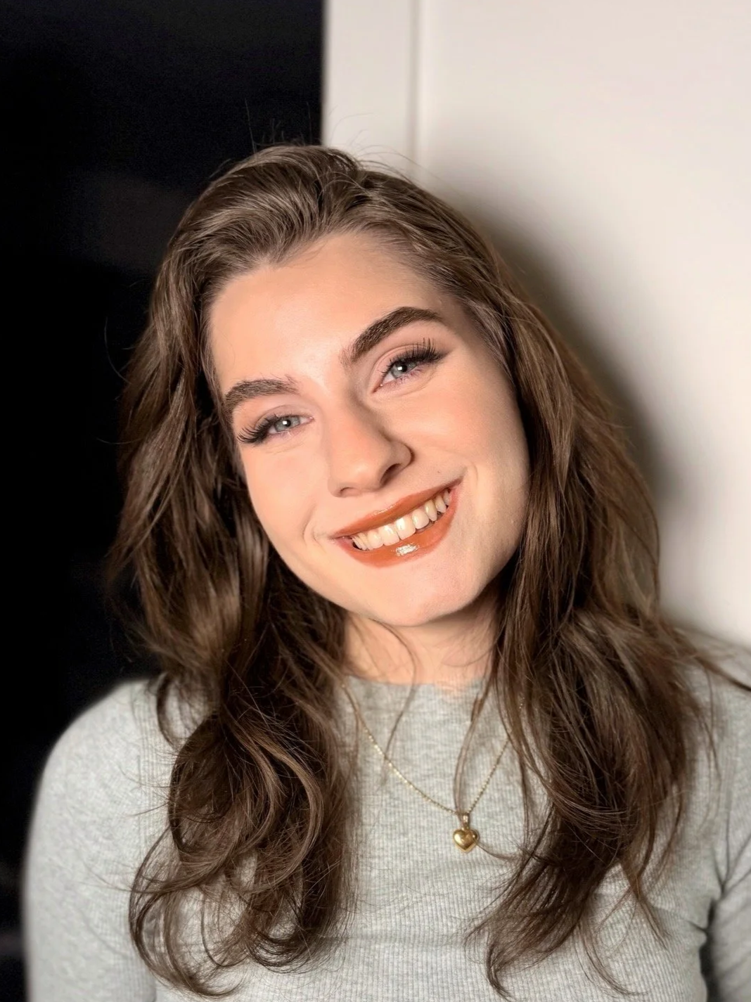 Close-up of a smiling woman with wavy brown hair, wearing a light gray top and gold heart-shaped necklace, against a black and white background.
