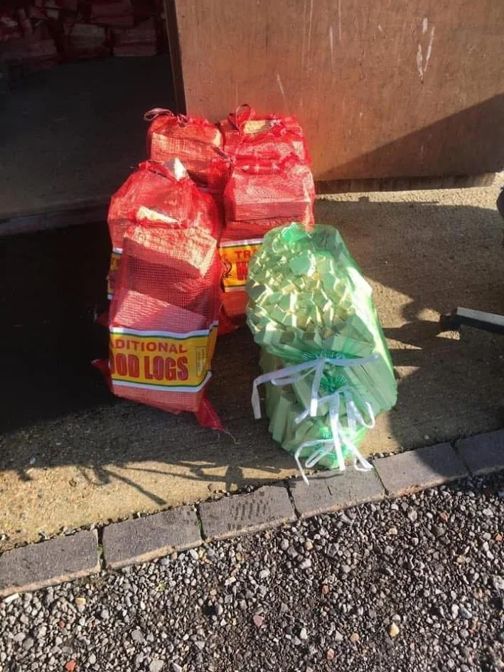 Bags of logs labeled 'Traditional Wood Logs' and a green bag tied with white ribbon, placed outside on gravel next to a wooden structure.