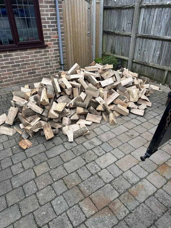 A pile of chopped firewood stacked on a brick patio near a wooden fence and house wall.