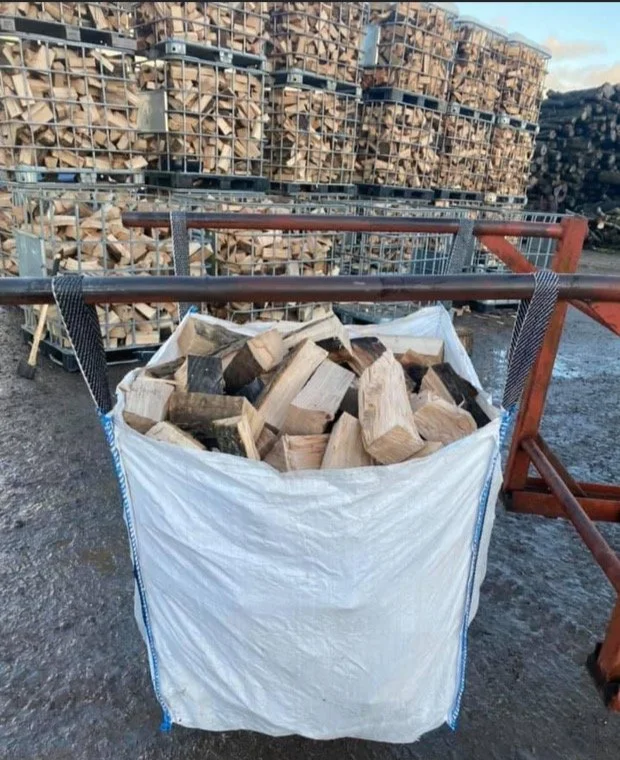 A large white bag filled with chopped firewood, standing outdoors on a dirt surface, with stacks of firewood in metal racks in the background.