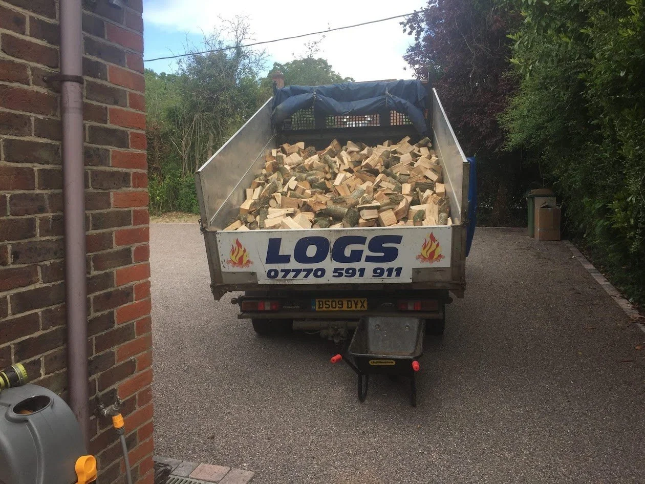 A truck filled with chopped firewood parked on a gravel driveway next to a brick wall and greenery.
