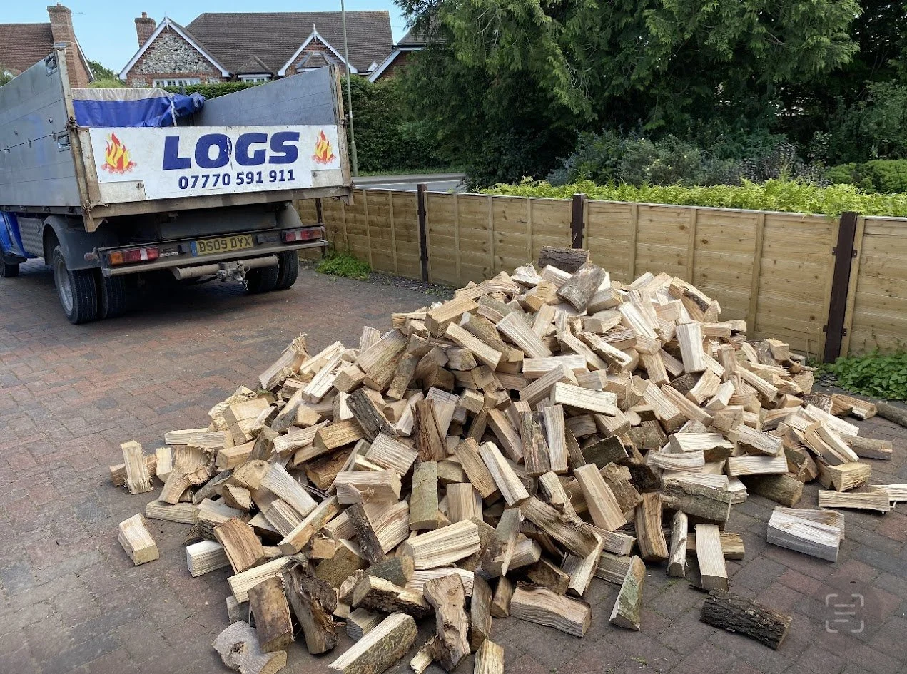 A pile of chopped firewood on a brick driveway next to a wooden fence, with a truck labeled 'LOGS' parked nearby.
