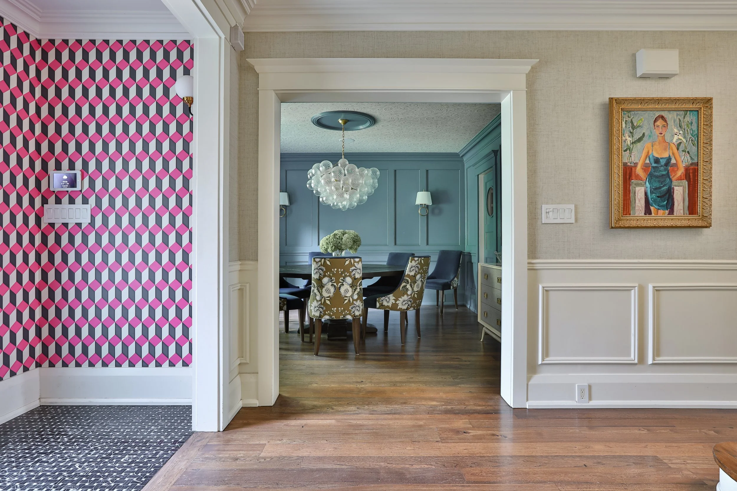 Image of a dining area with blue paneled walls, a round table with floral chairs, and wallpaper on the ceiling. The room has a mix of traditional decor and modern elements, with artwork on the walls and a sideboard visible.