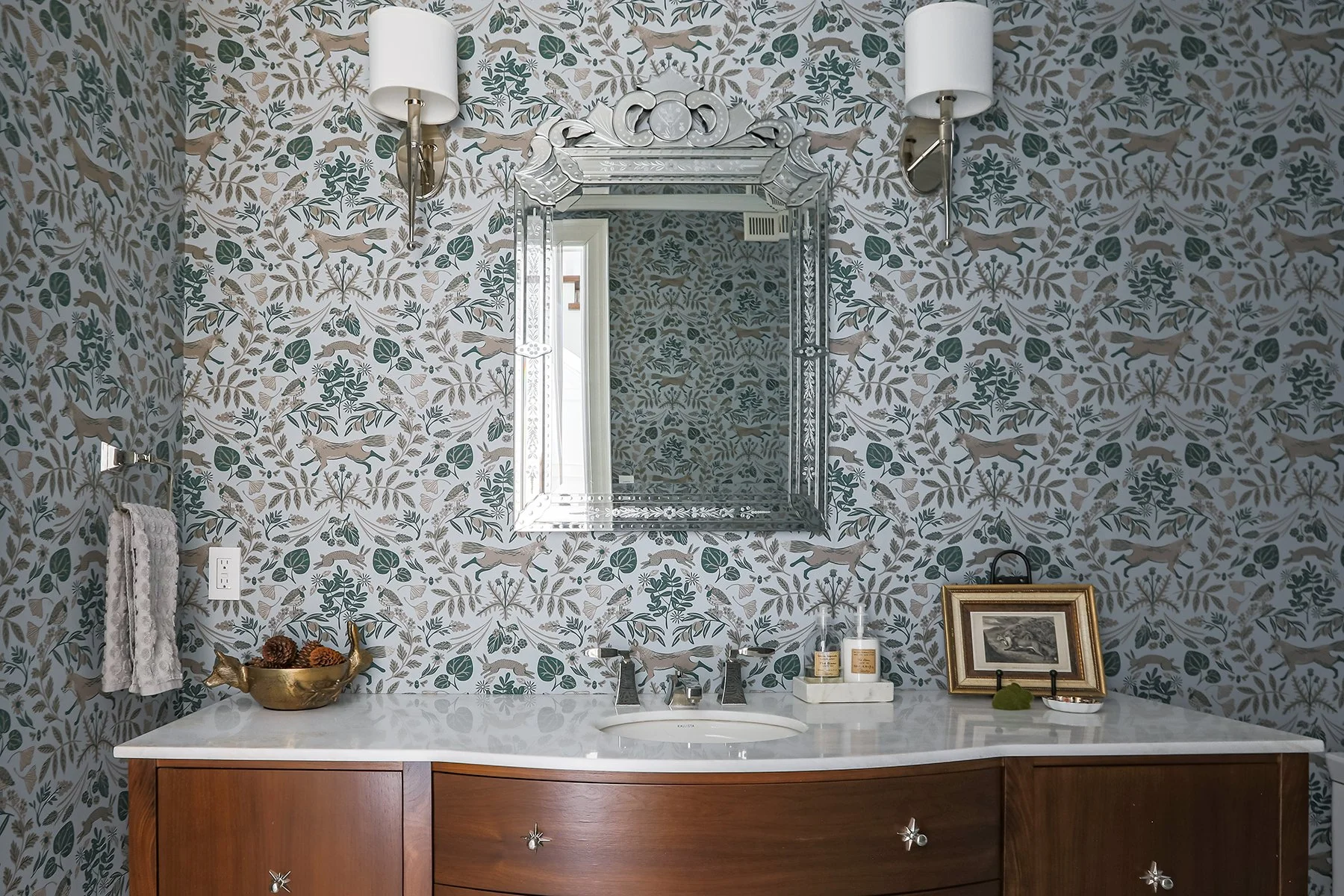 Bathroom with patterned wallpaper, mounted mirror, white sink with a wooden cabinet, and decorative items including framed photo, and jewelry dish.