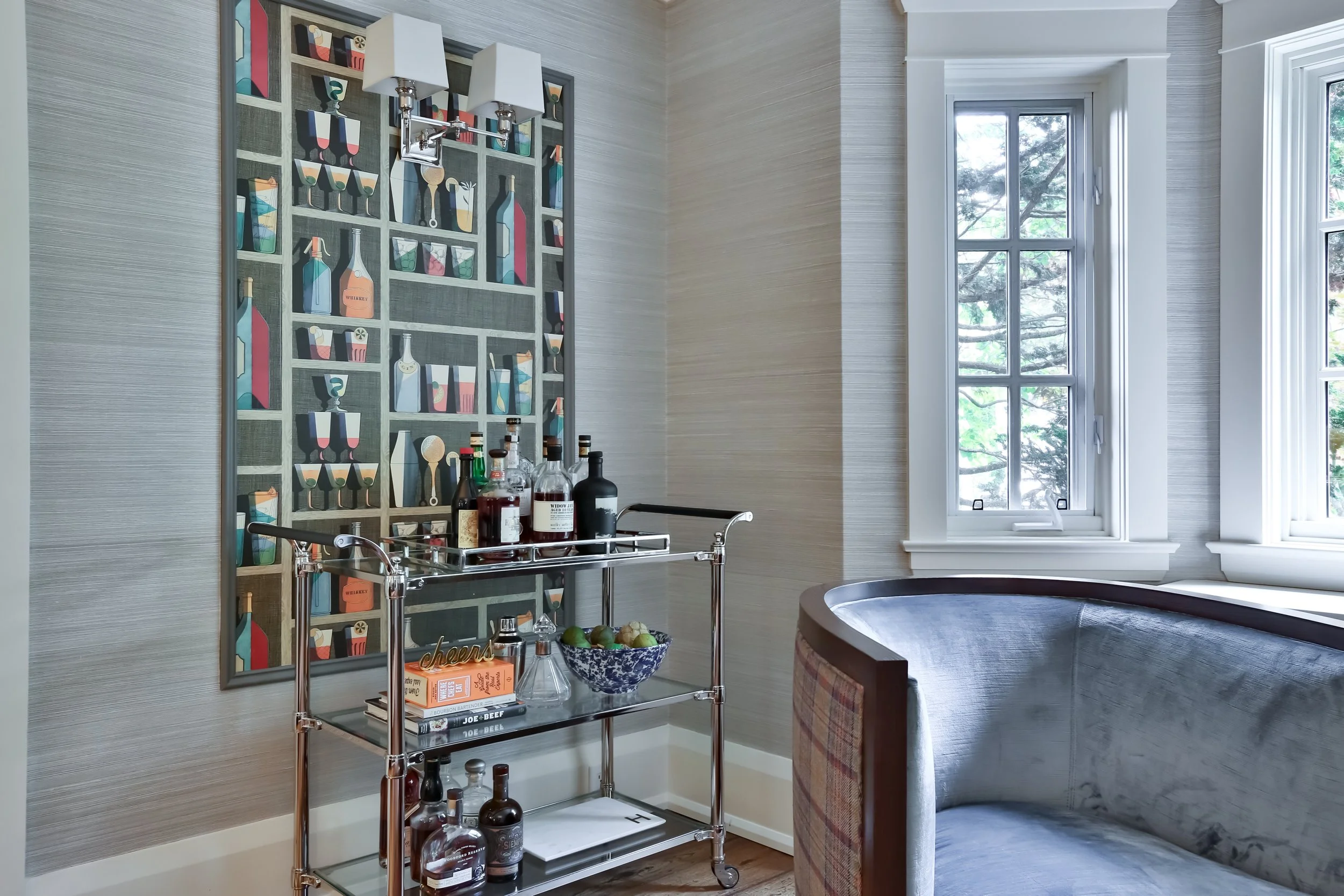 A corner of a room with large windows, a curved couch, wallpaper as framed art featuring colorful bottles and glasses, and a metal bar cart holding bottles, books, and a bowl of limes.