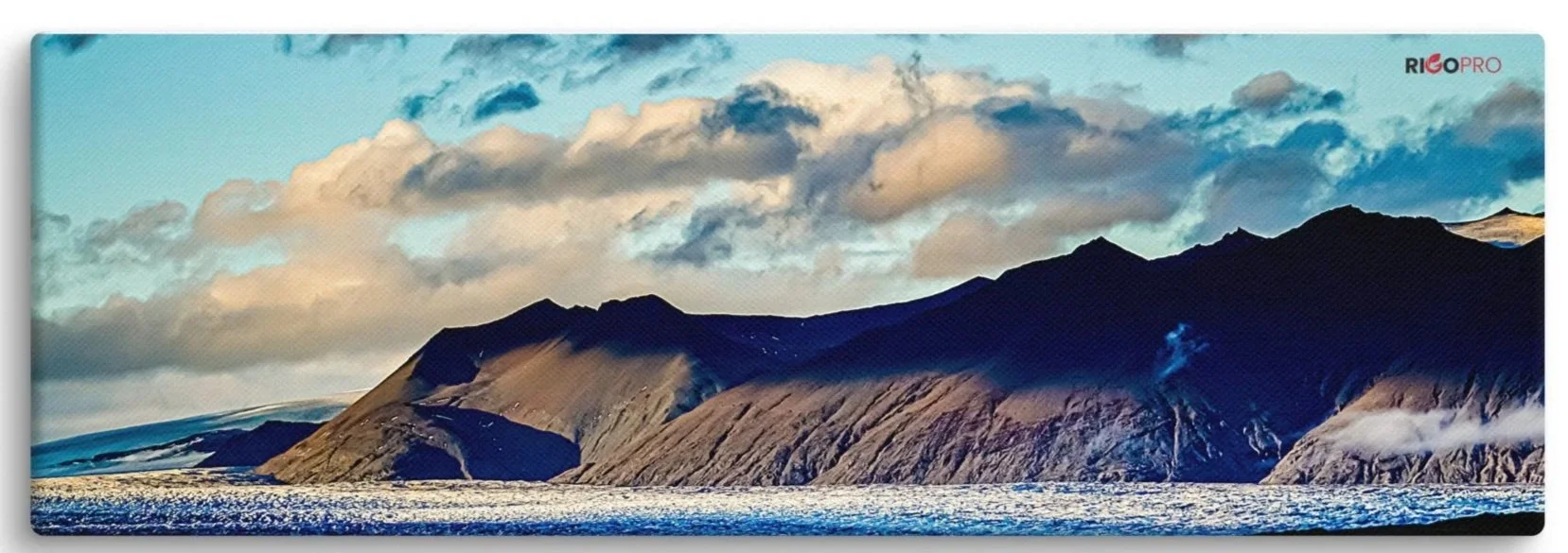 A long horizontal canvas print of a shadowy mountain flowing down to an icy glacier surface. The light blue sky is partly covered by clouds varying from fluffy white to shades of dark blue.