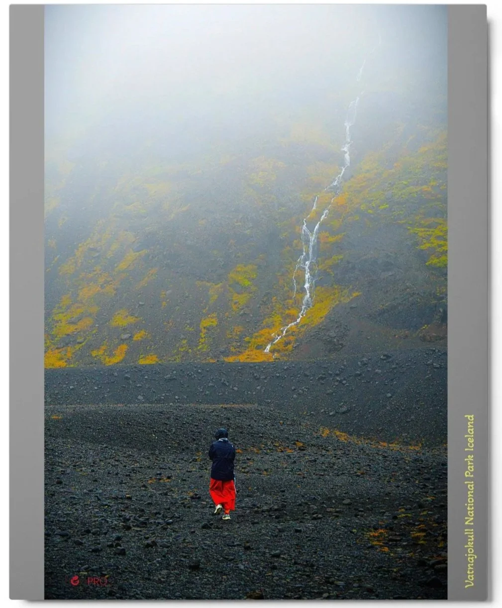 A rectangular color metal photograph of a lone woman wearing a bright red skirt, bundled in a black coat walking over dark volcanic rocky ground. The sky above is misty white with a lone stream of water flowing down a hillside towards her.