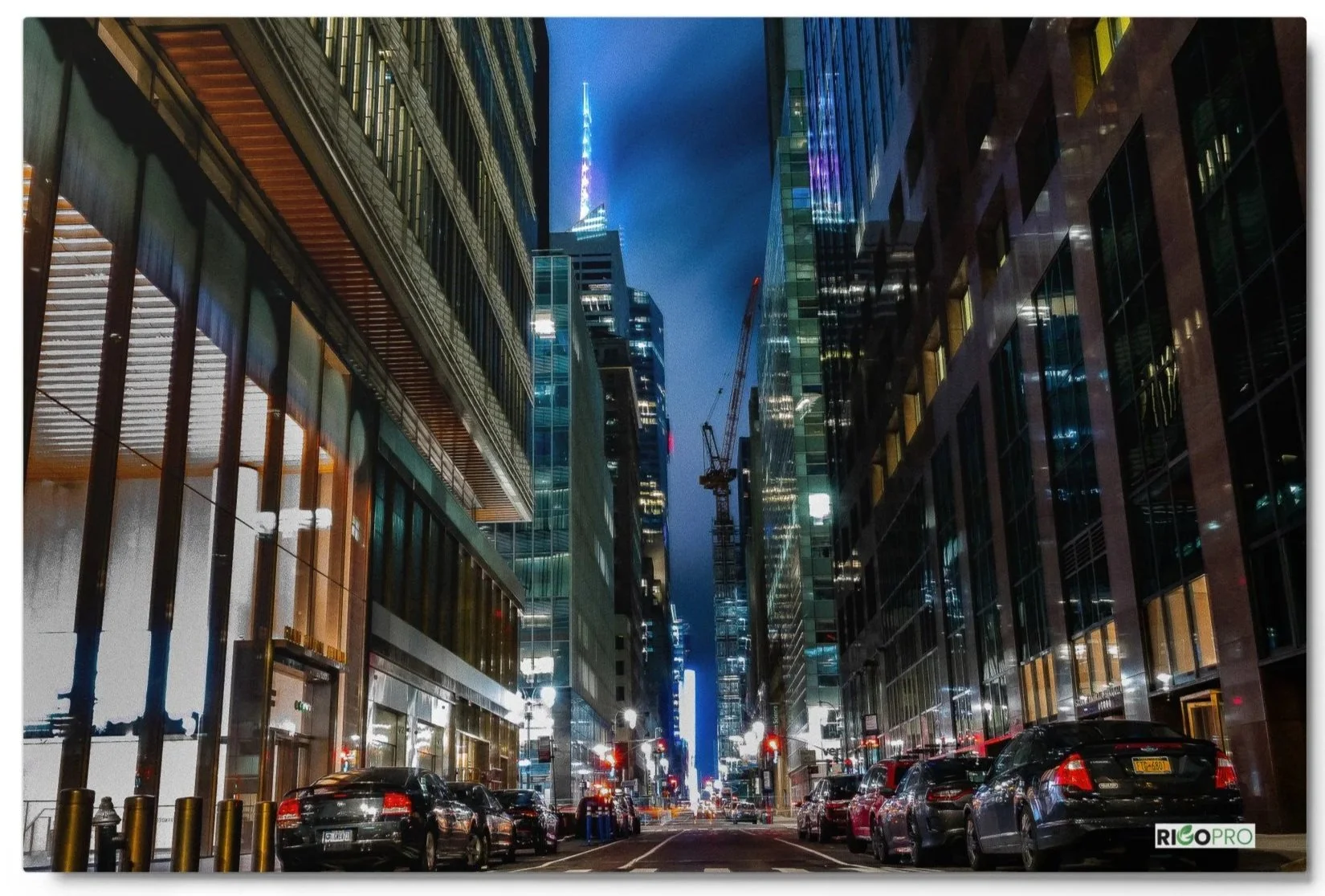 A glossy metal print photograph of a New York City street (43rd Street) shot at night from a low angle facing towards Times Square. The street is lined with cars and shiny building surfaces, reflecting the distant light amidst a misty steel-blue sky.