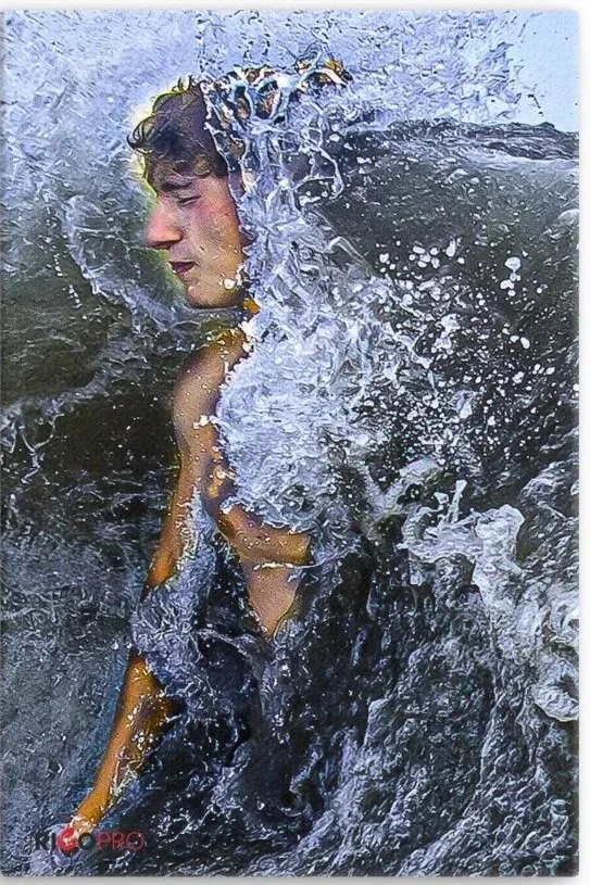 A closeup of a young man with eyes closed and pursed lips, emerging from a turbulent wave that is splashing against him with water drops flying in all directions.