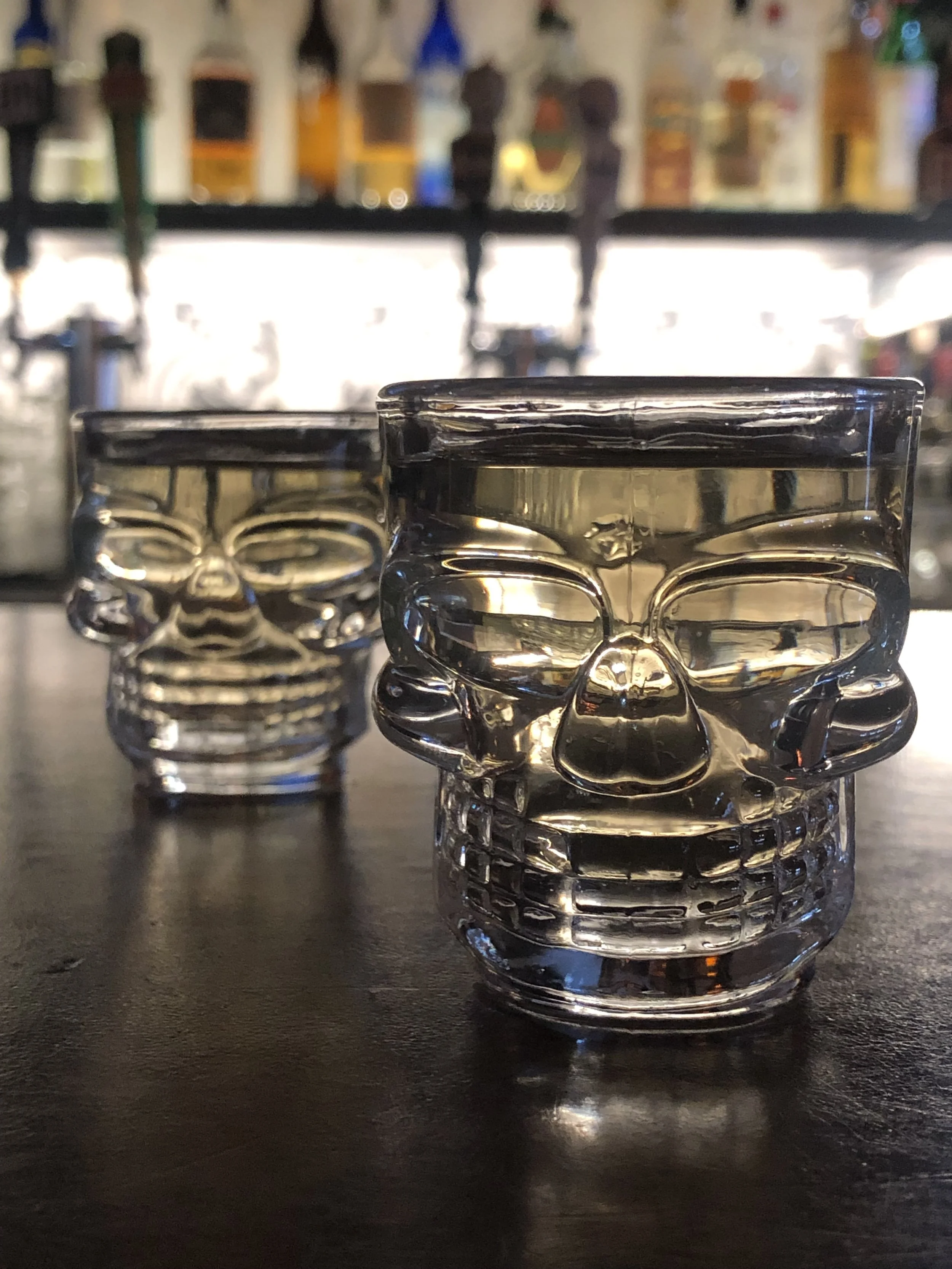 Two glass skull-shaped drinking glasses on a dark table, with a blurred bar background.