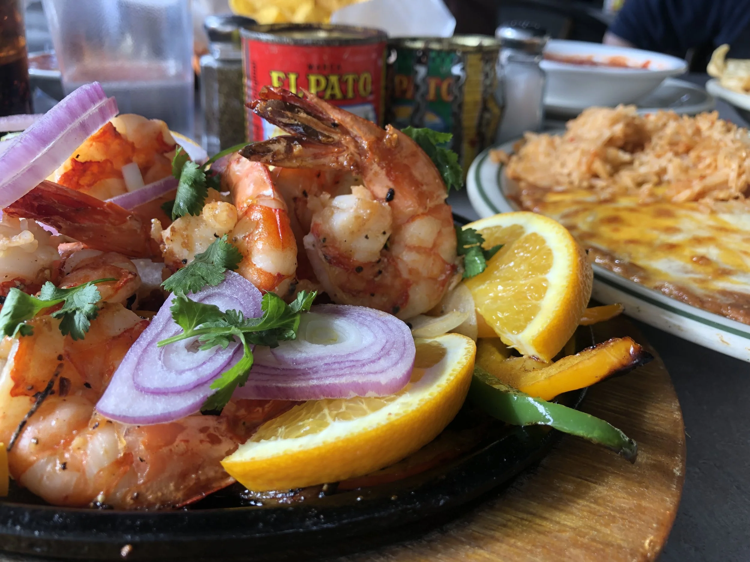 Close-up of a seafood dish with shrimp, garnished with cilantro, red onion slices, and lemon wedges, served on a hot skillet. In the background, there are plates of Mexican food, including rice and cheese-covered dishes, with canned tomatoes labeled 'El Plátano' and 'Ticos' also visible.