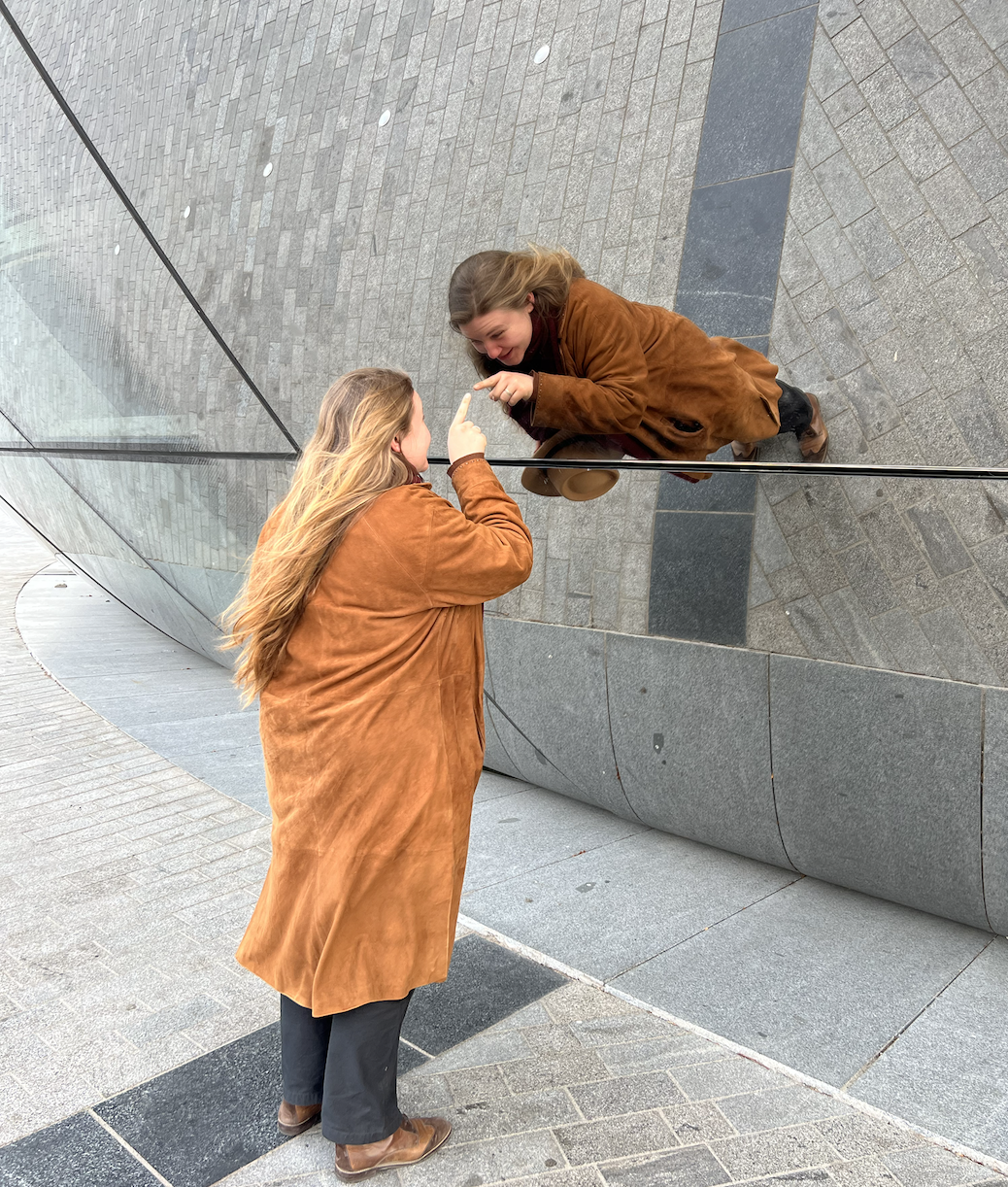 A woman with a brown coat mirrored in a building with reflective glass, gesturing with her hand. Manuela Zahner.