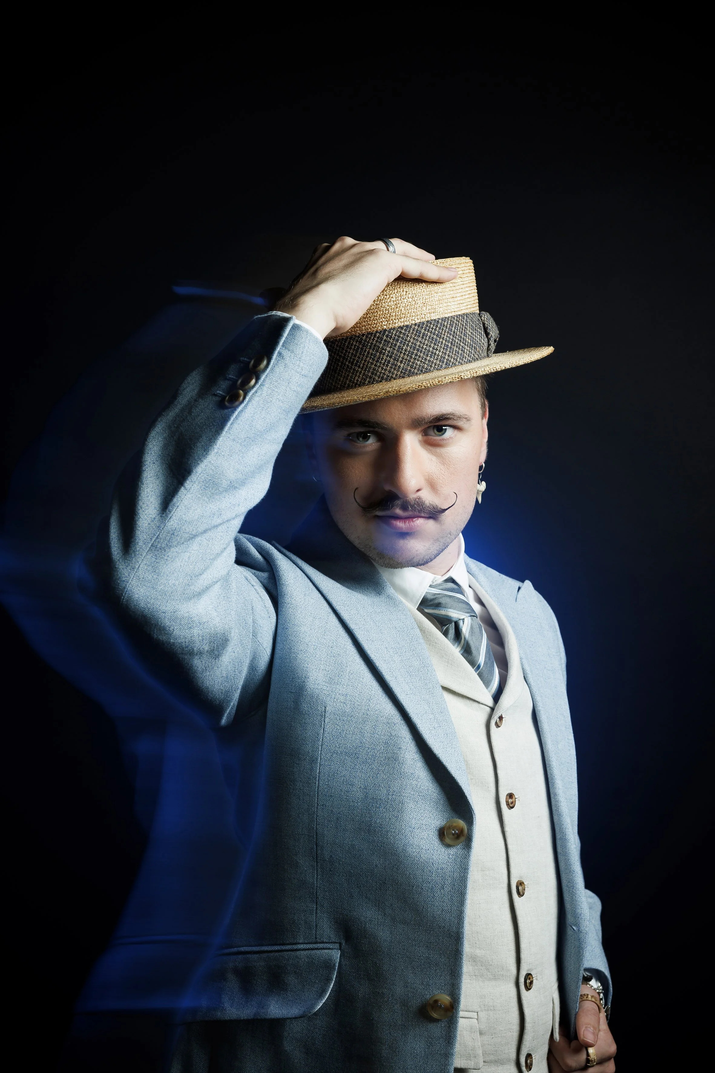 A young man in vintage attire holding a straw fedora hat, looking confidently into the camera against a dark background. Lukas Hefti.