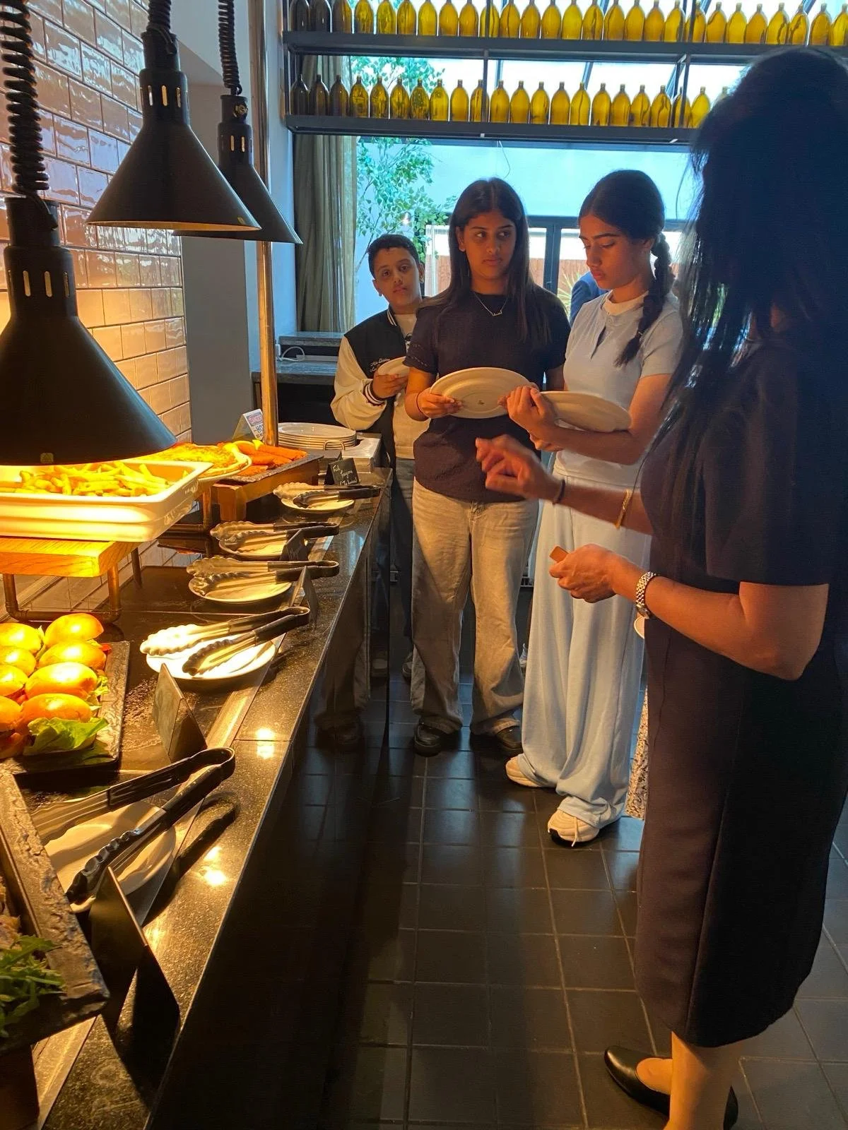 People standing in line at a buffet station, selecting food from a tray of French fries, with plates in hand, in a restaurant or buffet setting.
