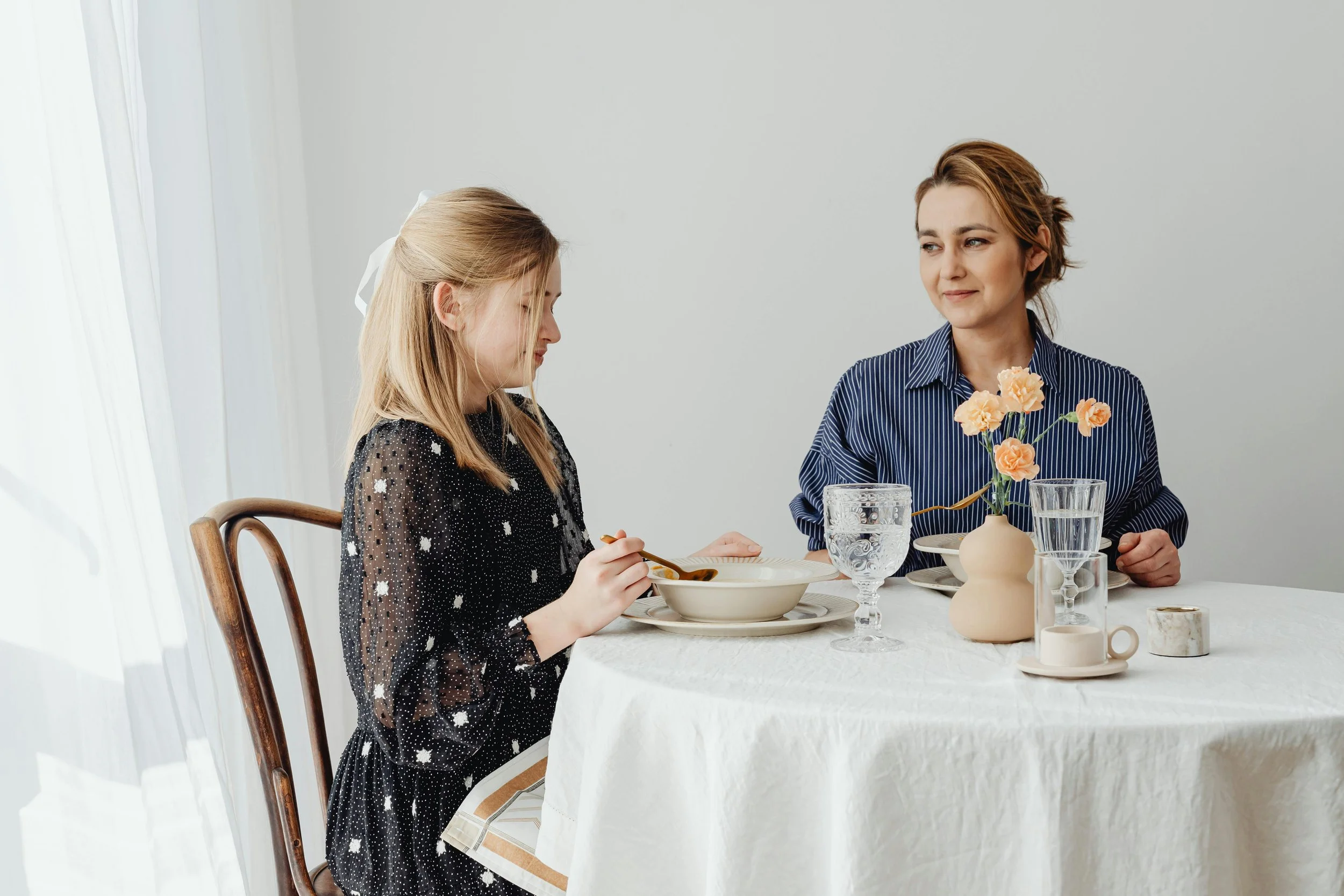 A woman and a young girl sitting at a table having a meal, with a white background and decorated with vases and glasses.