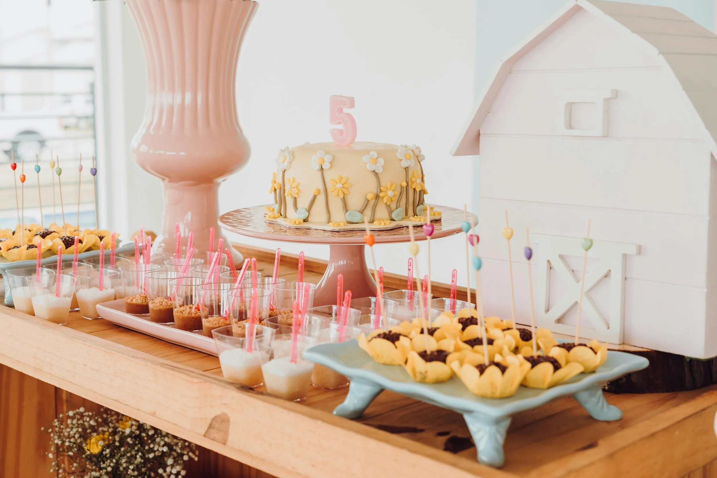 A dessert table with a pink cake topped with a "5" candle and flower decorations, surrounded by mini desserts and colorful candies, against a pastel background.