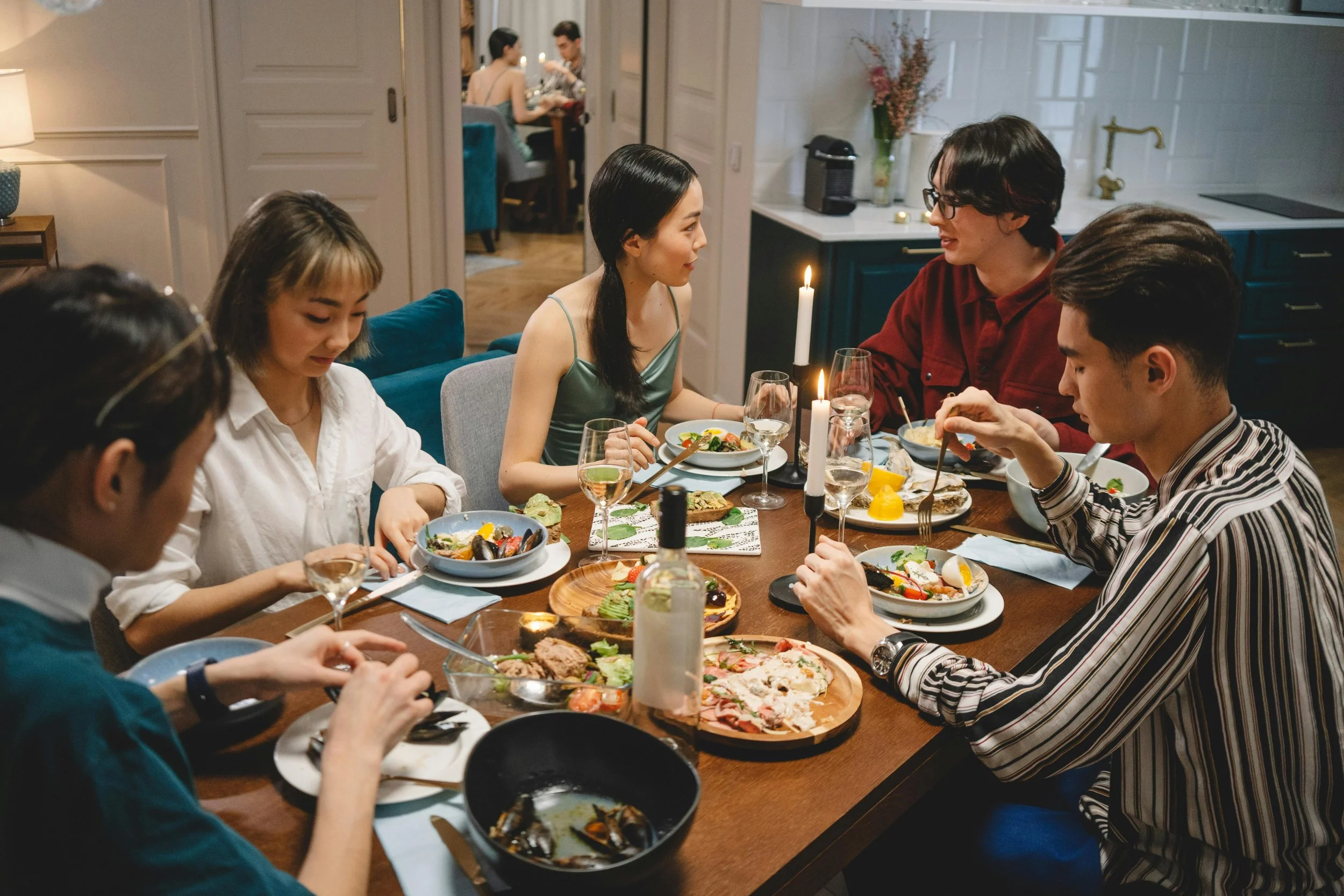 Group of six friends enjoying a dinner party at a table with various dishes, candles, and glasses of wine in a cozy home setting.