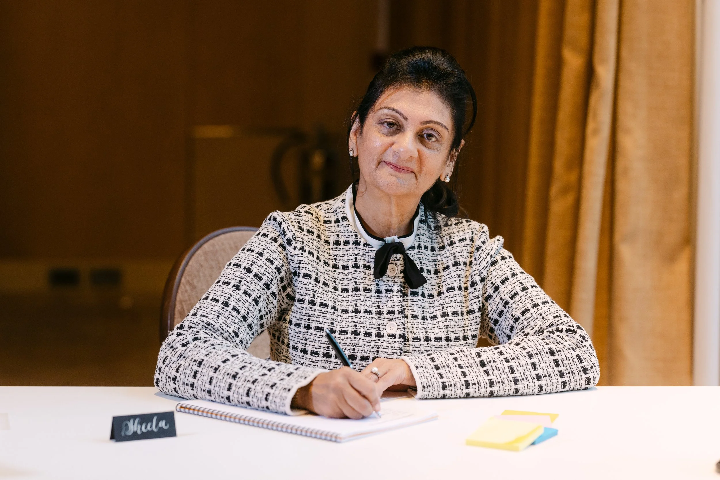 A woman sitting at a table, writing on a notepad, with a nameplate in front of her that reads 'Meda'. She is wearing a black and white patterned blazer with a black ribbon around her neck, and has dark hair pulled back. The background includes a wooden wall and beige curtains.