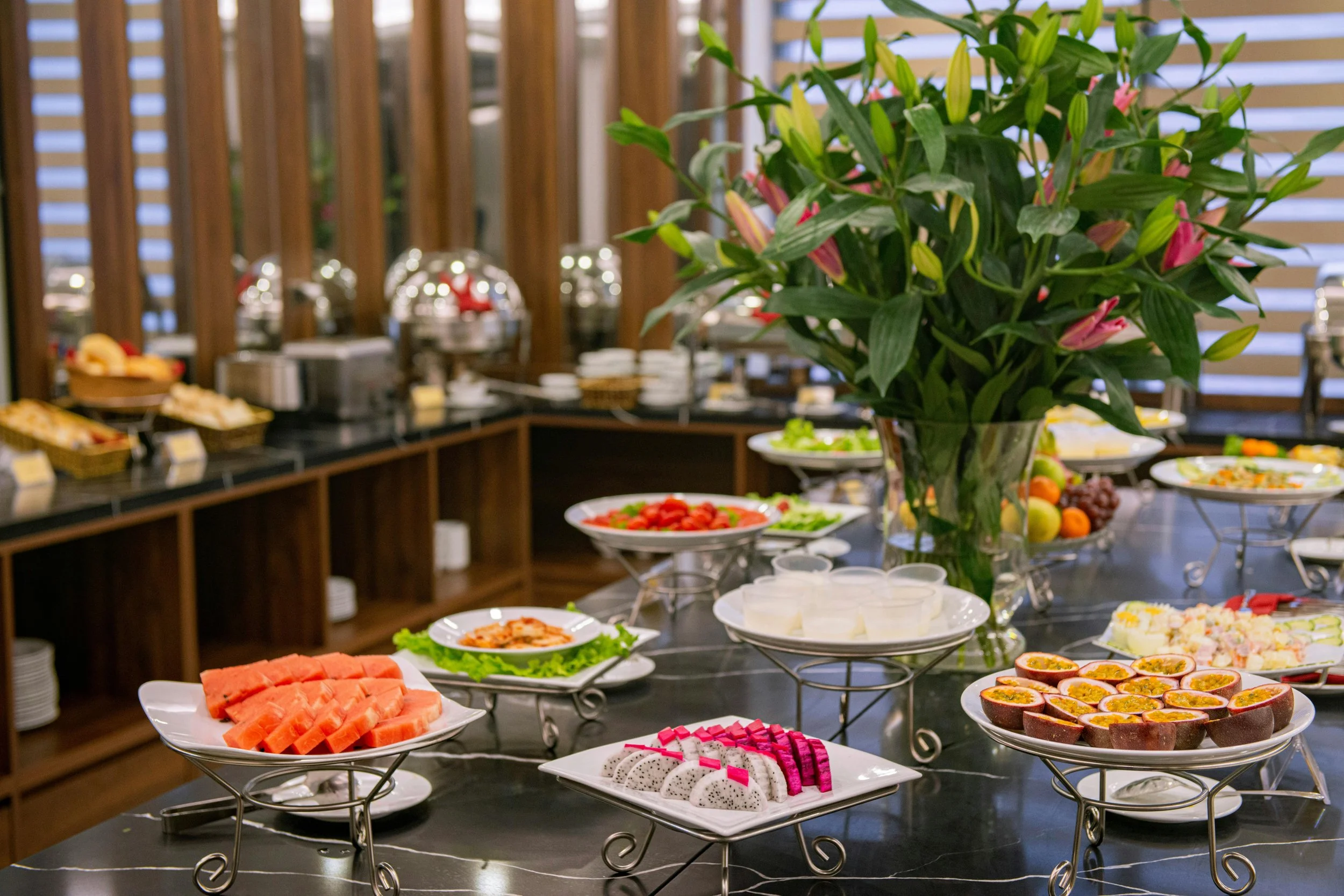 Buffet table with various fruits, salads, and desserts, decorated with a large bouquet of lilies in a glass vase.