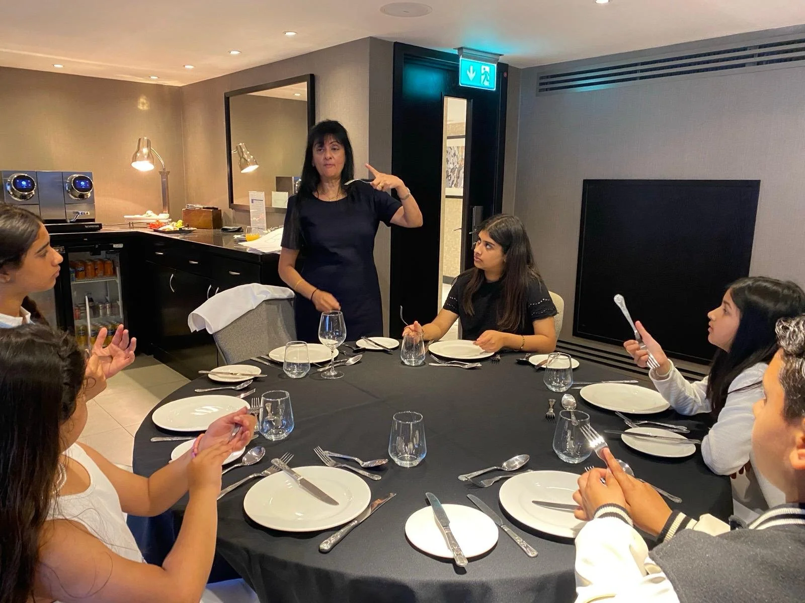 A woman standing at a round dining table with six seated women, gesturing with her right hand, in a restaurant setting with table settings, glasses, and a small buffet area in the background.