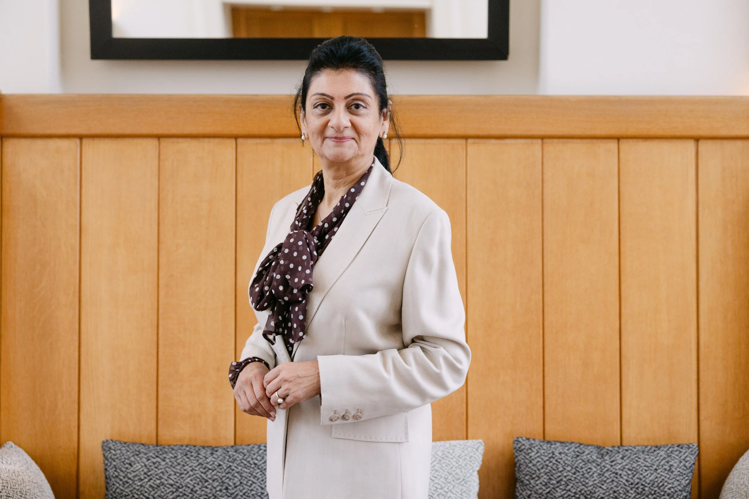 A woman dressed in a white suit with a polka dot scarf, standing indoors in front of a wooden paneled wall and a mirror.