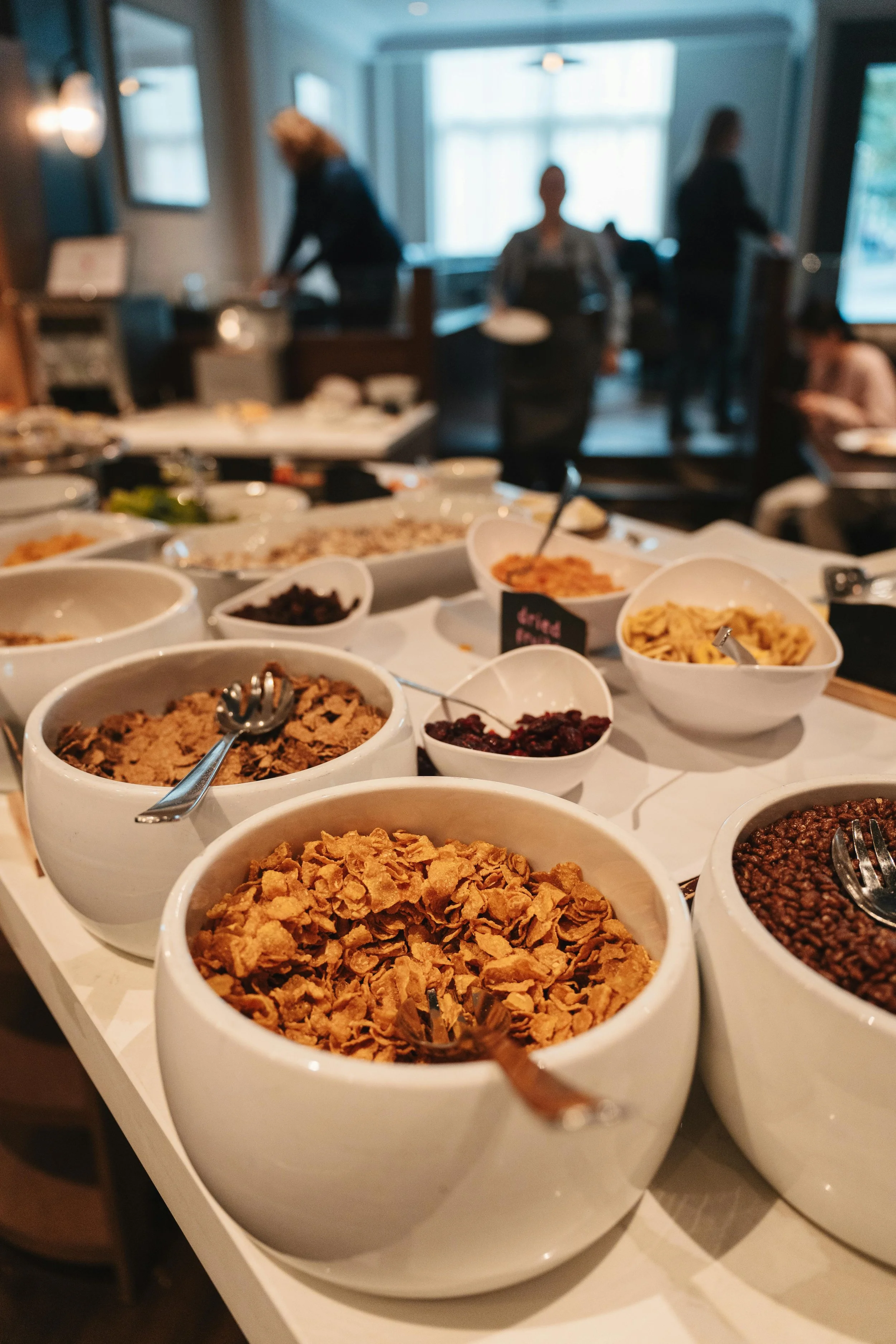 Bowls of cereal, dried fruit, and toppings on a buffet table at a restaurant.