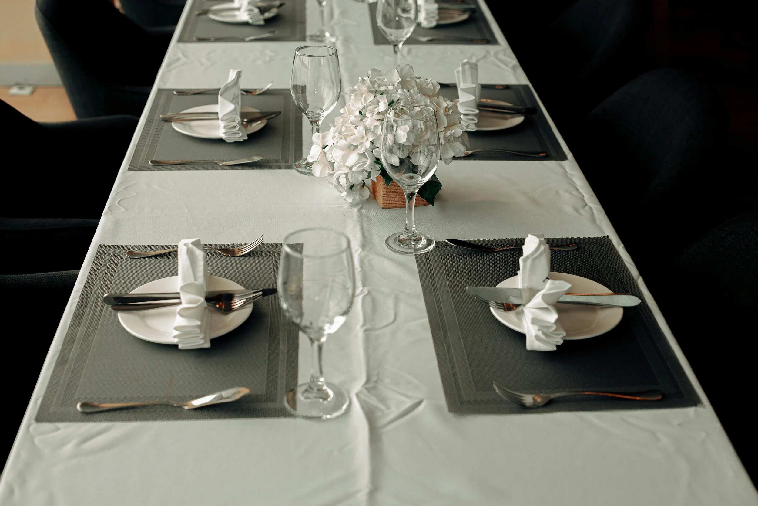 A formal dining table set with white tablecloth, gray place mats, white napkins, silverware, empty wine glasses, and a floral centerpiece of white hydrangeas.