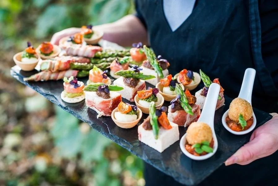 Person holding a black slate serving platter with various gourmet appetizers, including mini tartlets, canapés, and croquettes, garnished with vegetables and herbs.