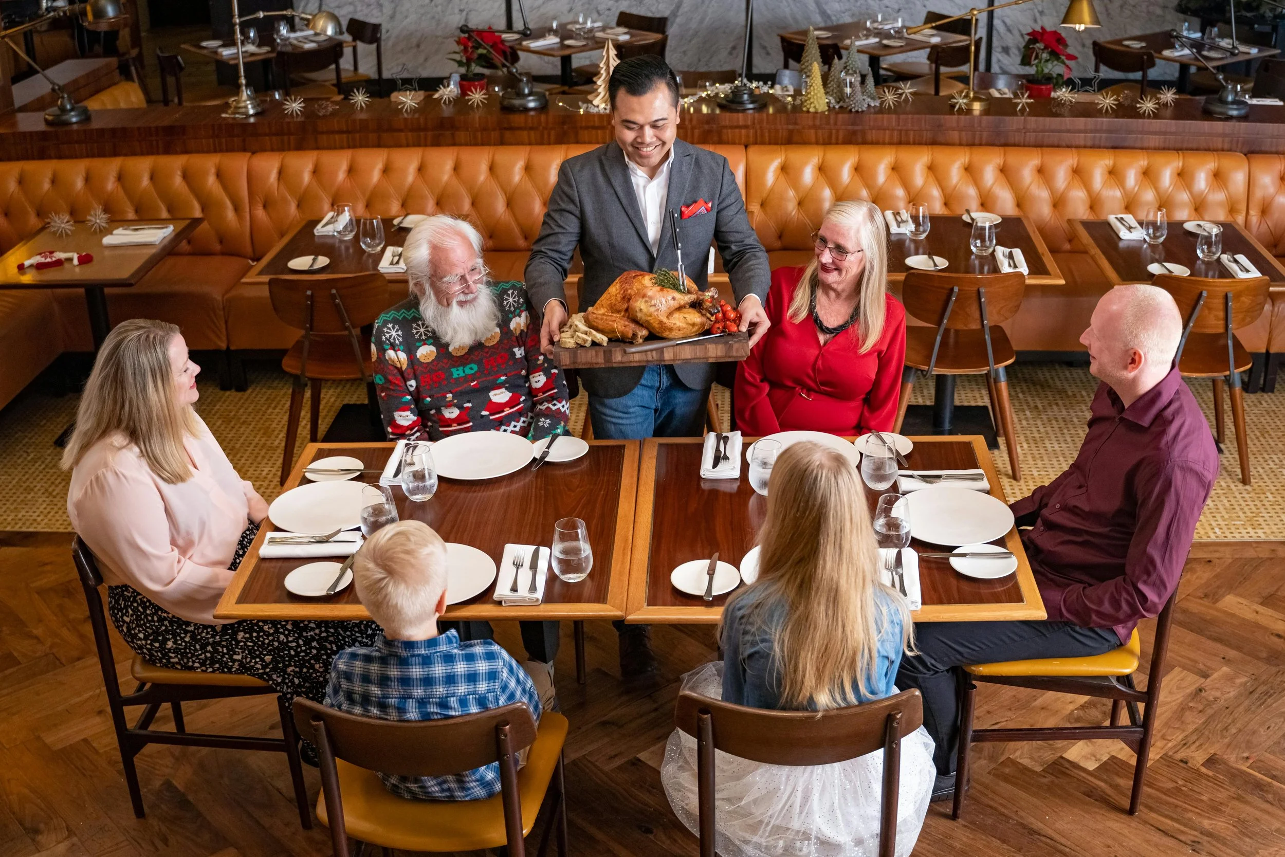 A group of seven people gathered at a restaurant table for a holiday celebration. A server is presenting a roasted turkey on a tray, smiling. The table is set with white plates, silverware, and glasses. The diners include older adults and children, w