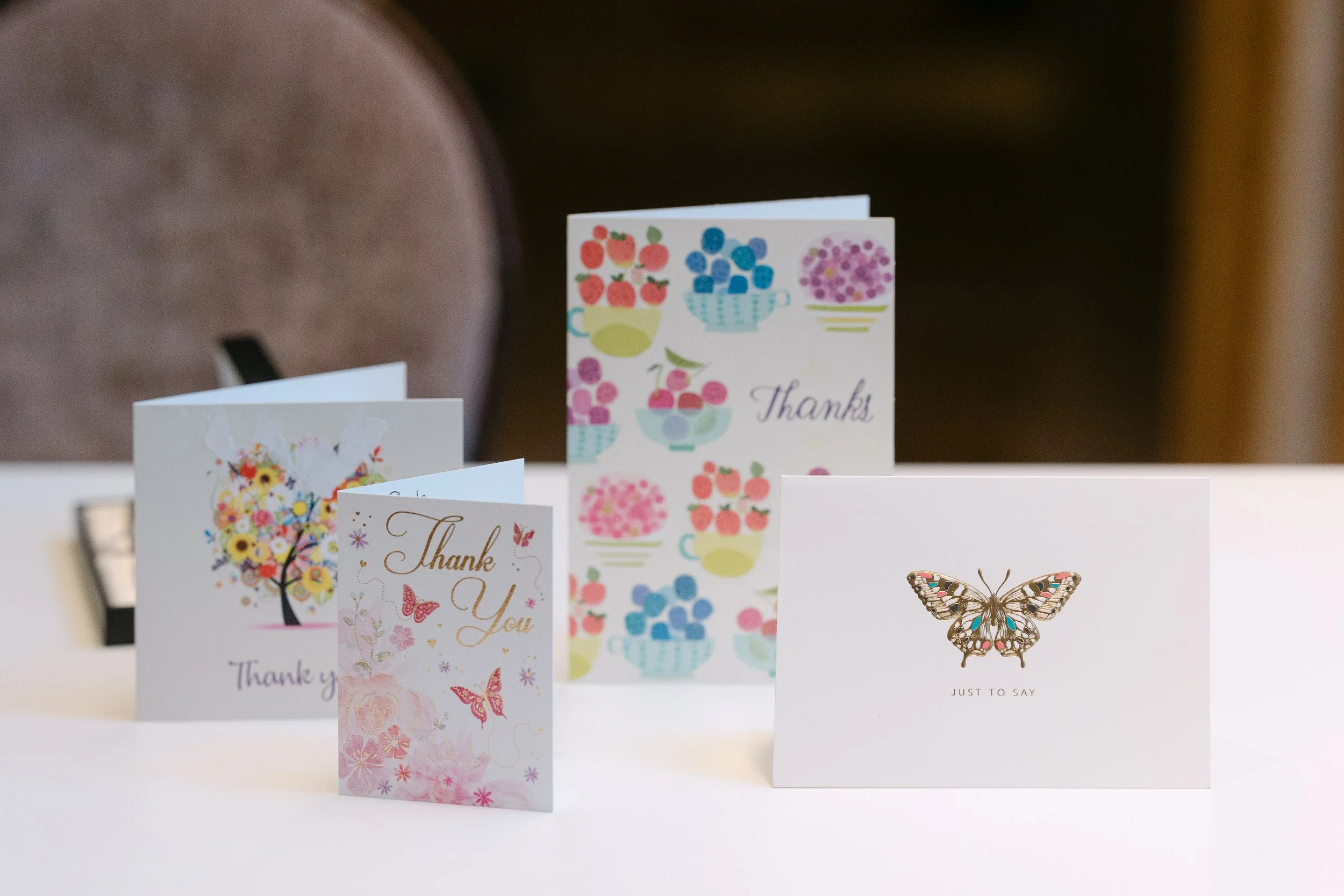 Four thank you cards with floral and butterfly designs on a white surface. The cards feature colorful illustrations, including flowers, a butterfly, and the word "Thanks."