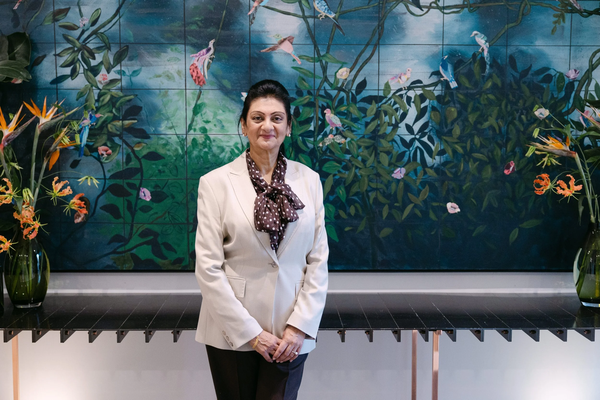 A woman in a beige blazer and dark pants standing in front of a large mural depicting a vibrant jungle scene with birds, plants, and flowers, flanked by two vases with tropical flowers.