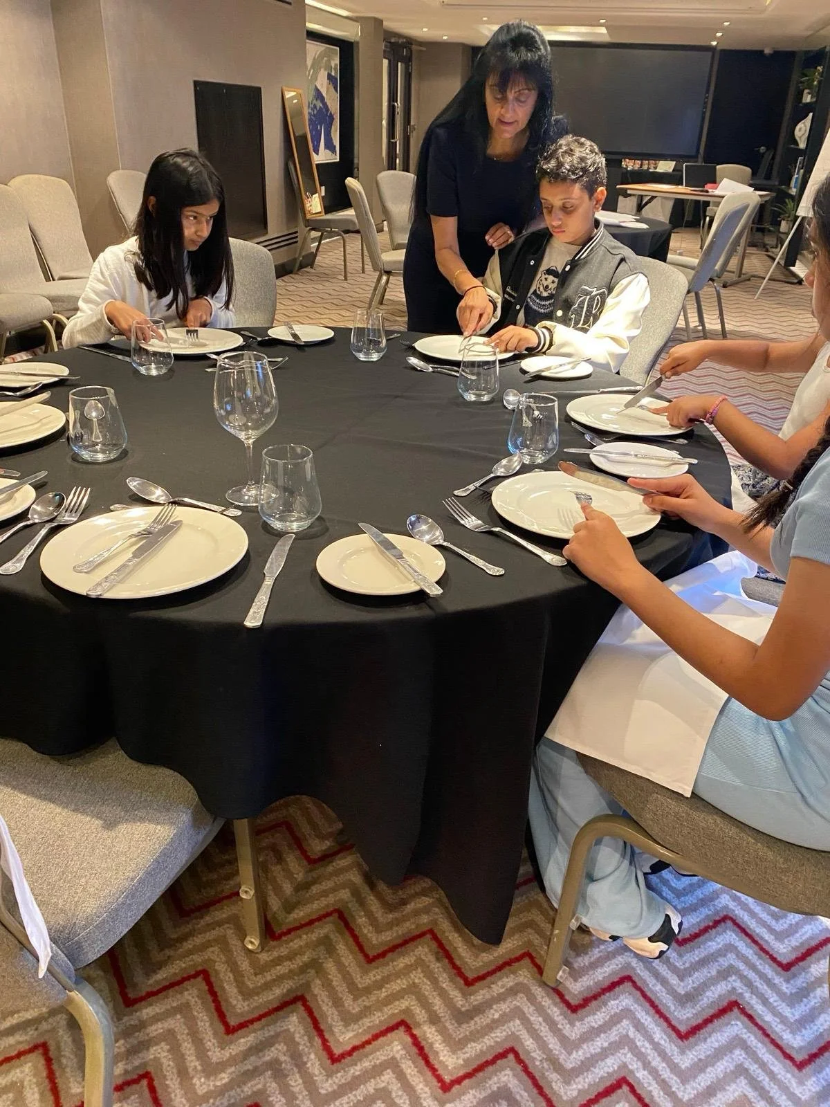 A woman is helping a young boy set the table with plates, utensils, and glasses. Three other girls sit around the table in a well-lit room, preparing for a meal.