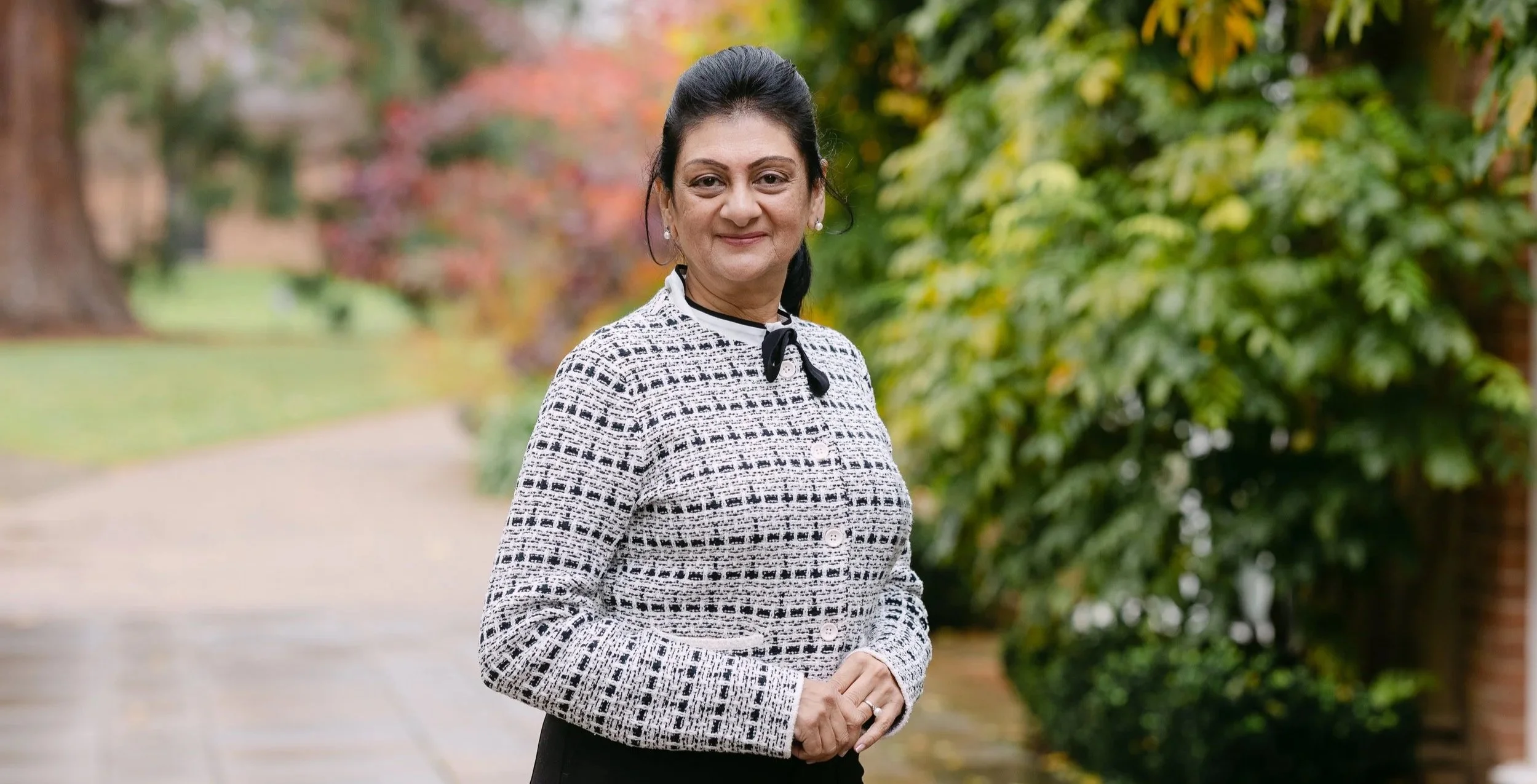 A woman in a patterned black and white blazer with a black bow tie stands outdoors on a pathway with autumn foliage in the background.