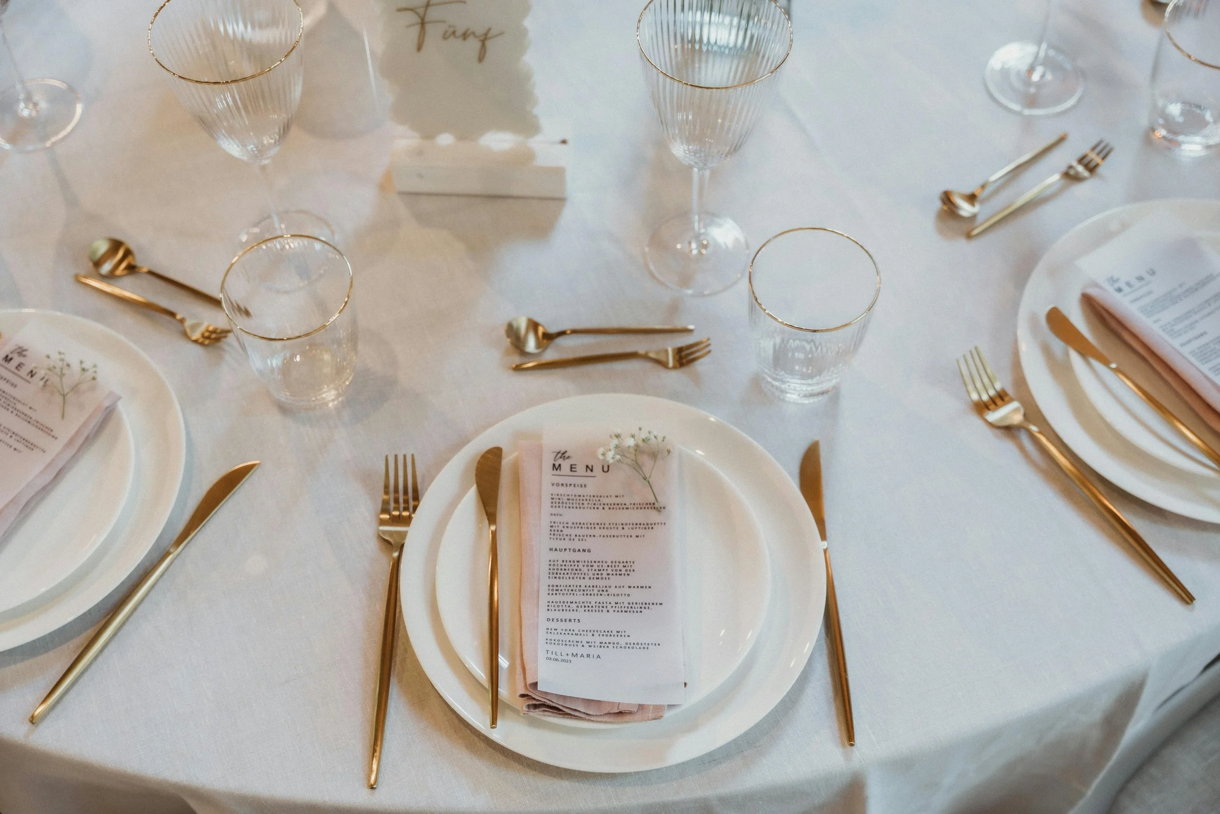 Elegant dining table set for a formal event, with white tablecloth, gold cutlery, clear glasses with gold rims, plates with pink napkins, and a menu card on each plate.