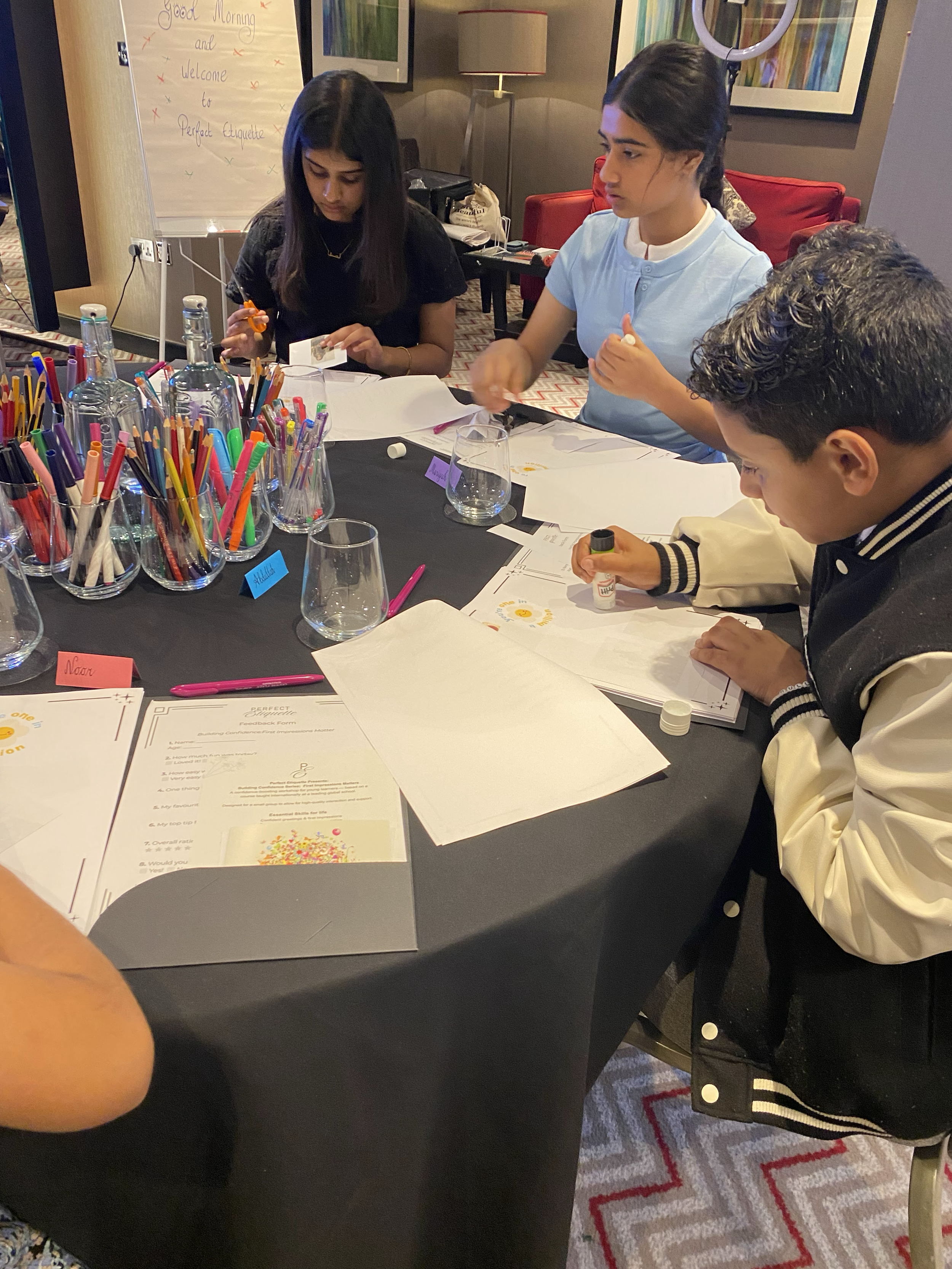 Three young individuals sitting at a black table with art supplies and papers, working on a craft activity in a room with framed artwork, a lamp, and a whiteboard that reads 'Good Morning and Welcome to Perfect Etiquette.'