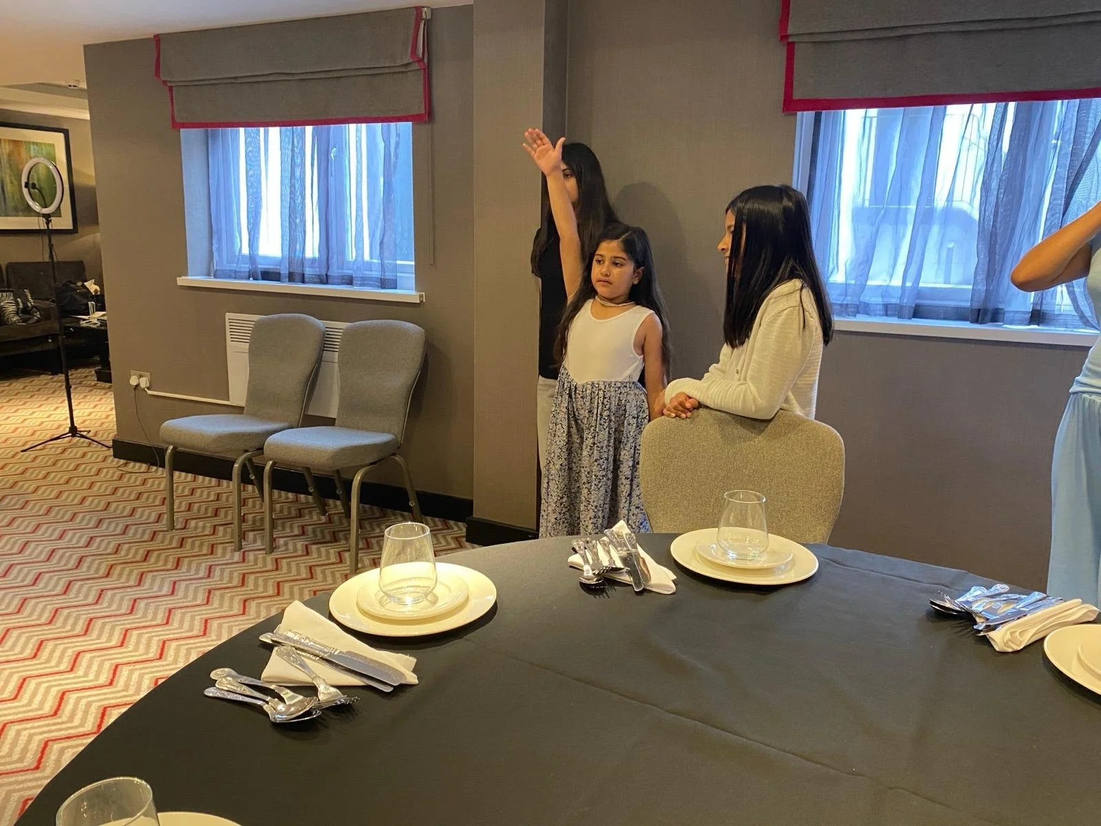Three women and a young girl standing near a wall in a dining room. The table in the foreground is set with plates, glasses, and utensils.