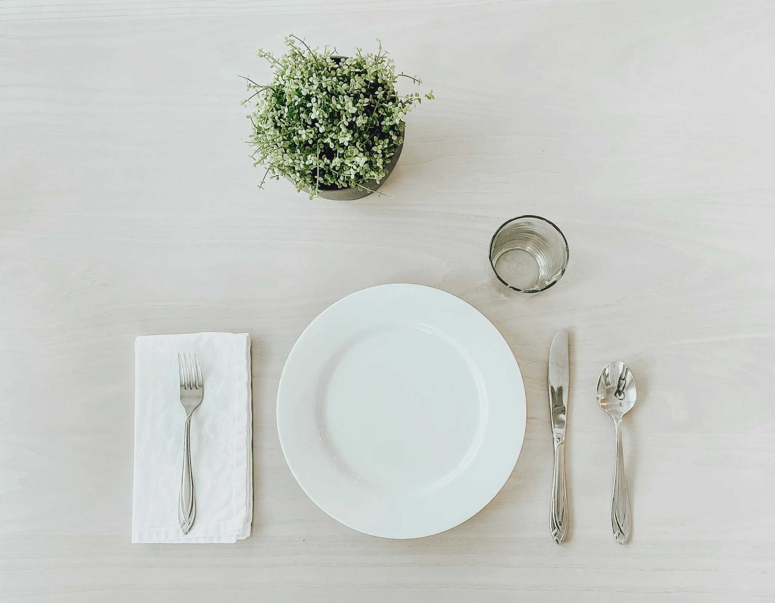 Empty white plate centered with a fork on a white napkin on the left, a knife on the right, and a spoon on the right of the plate. A glass of water is above the utensils, and a potted green plant is at the top left on a light-colored wooden table.