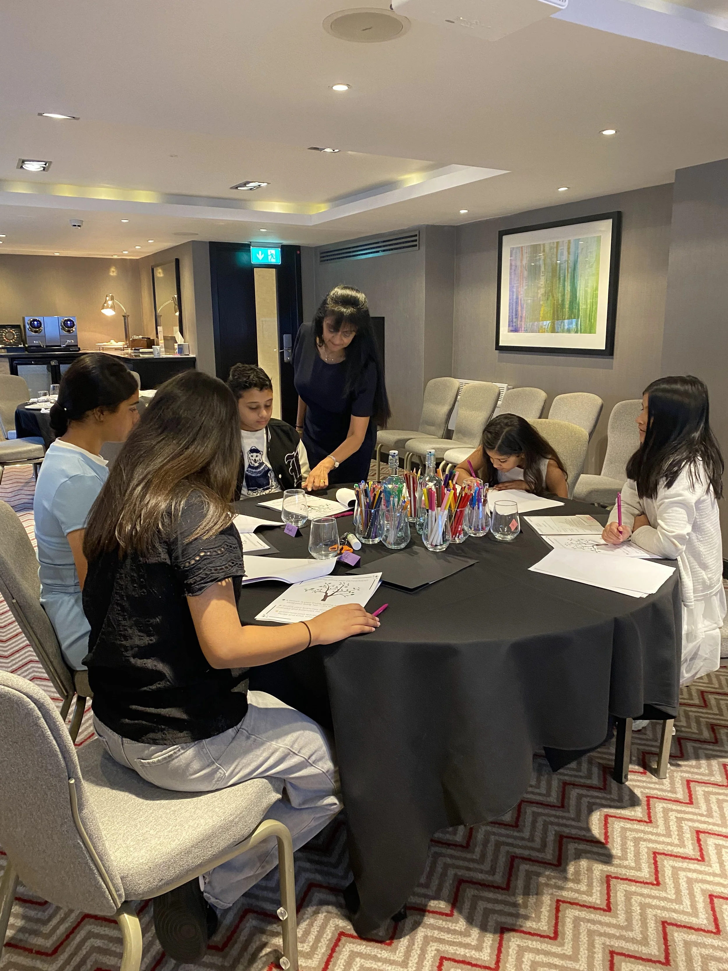 A group of young girls sitting around a round table with drawing materials, working on art projects, while an adult woman supervises in a well-lit indoor setting.