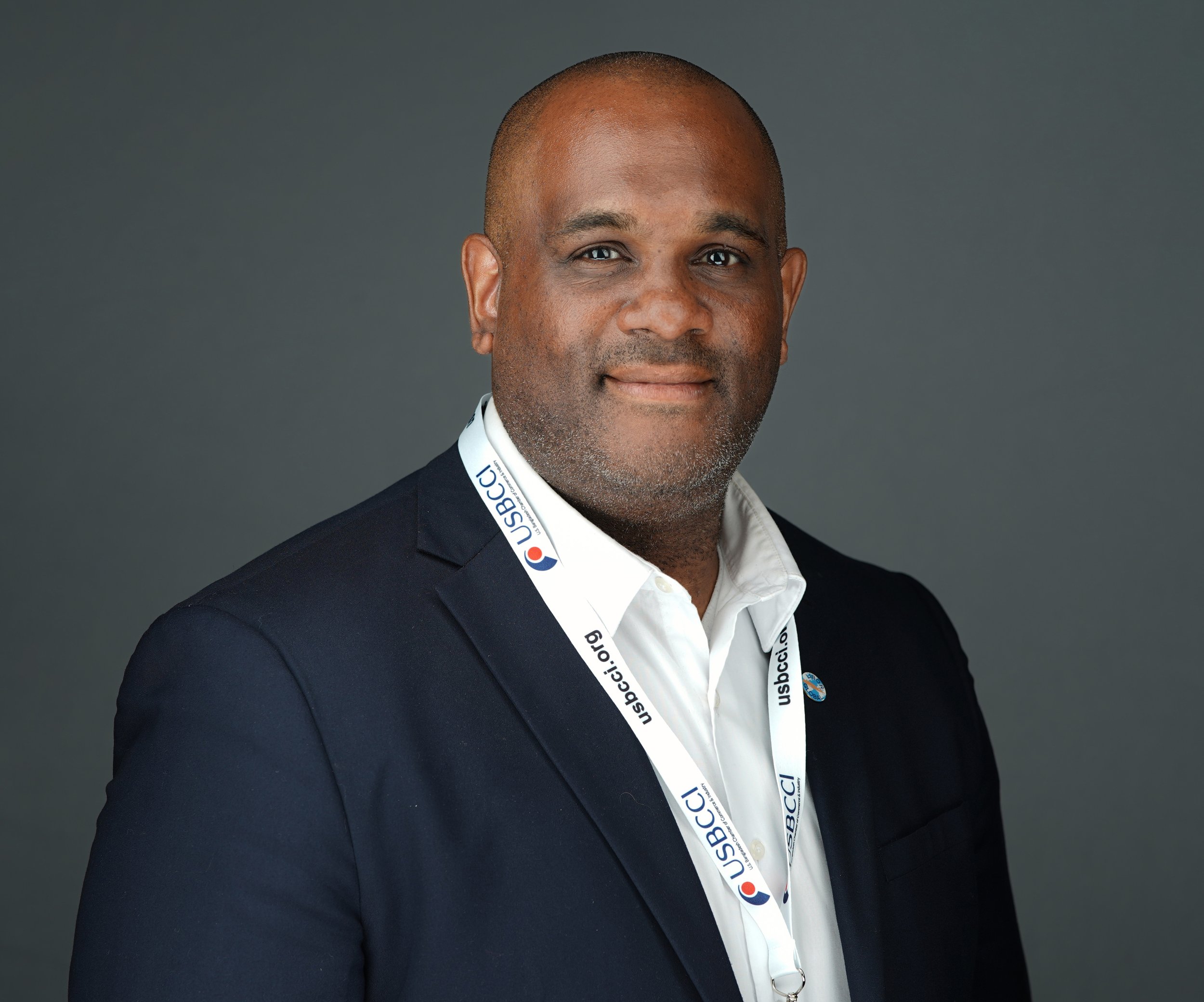 Portrait of a middle-aged African American man with short hair, wearing a dark suit, white shirt, and a lanyard with 'USBCC' written on it. He has a slight smile and is posed against a solid gray background.
