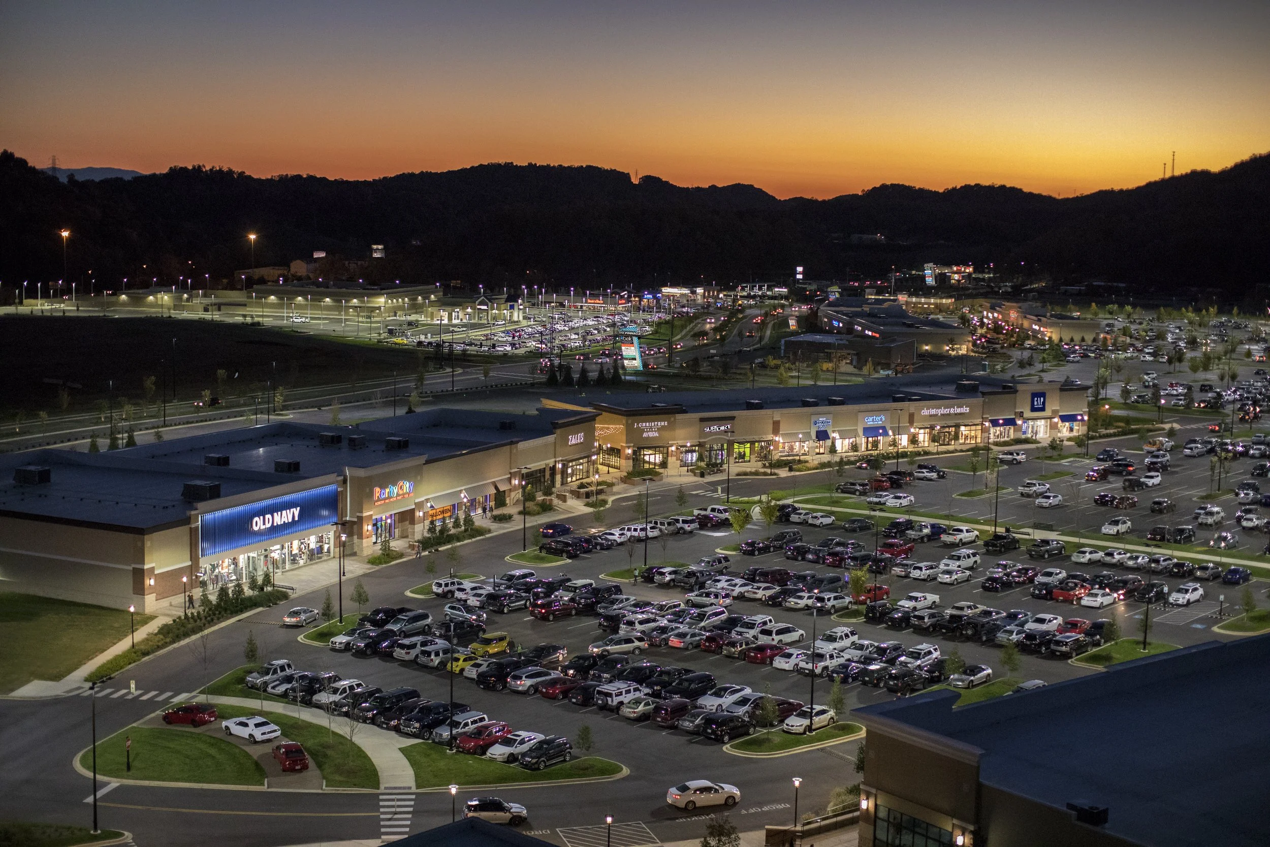 A large shopping mall parking lot during dusk with cars parked and streetlights on. Buildings with store signs are visible in the foreground, and hills with a colorful sunset sky are in the background.