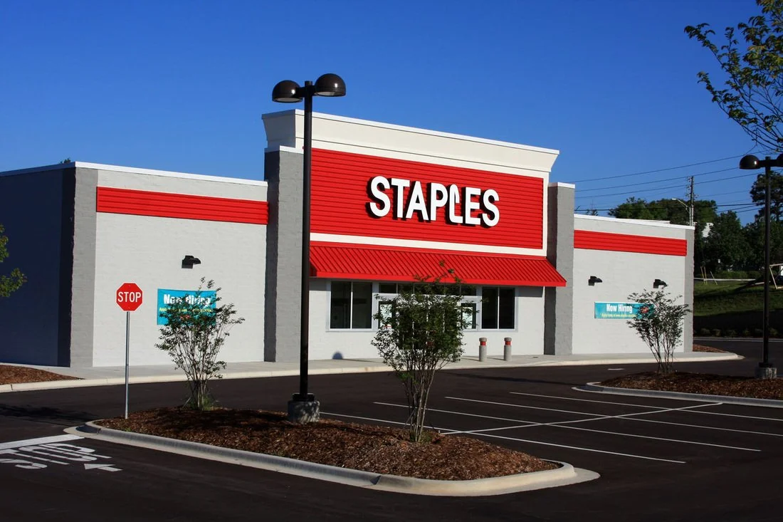 Empty parking lot in front of a Staples store with a large red sign, small trees, and a blue sky.