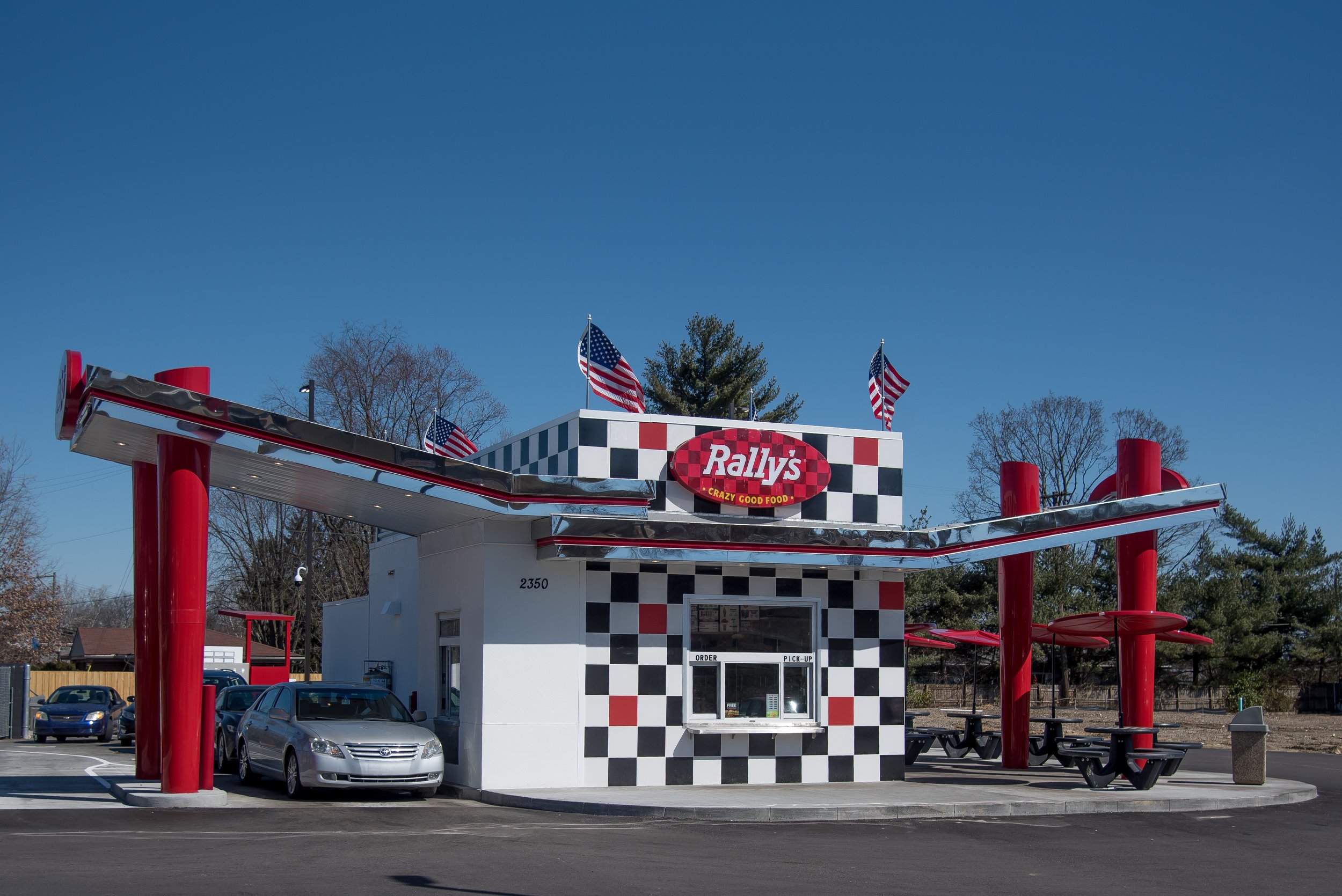 A fast-food restaurant named Rally's with a black and white checkered pattern and red accents. The building has a car drive-thru, American flags on top, and a small order window.