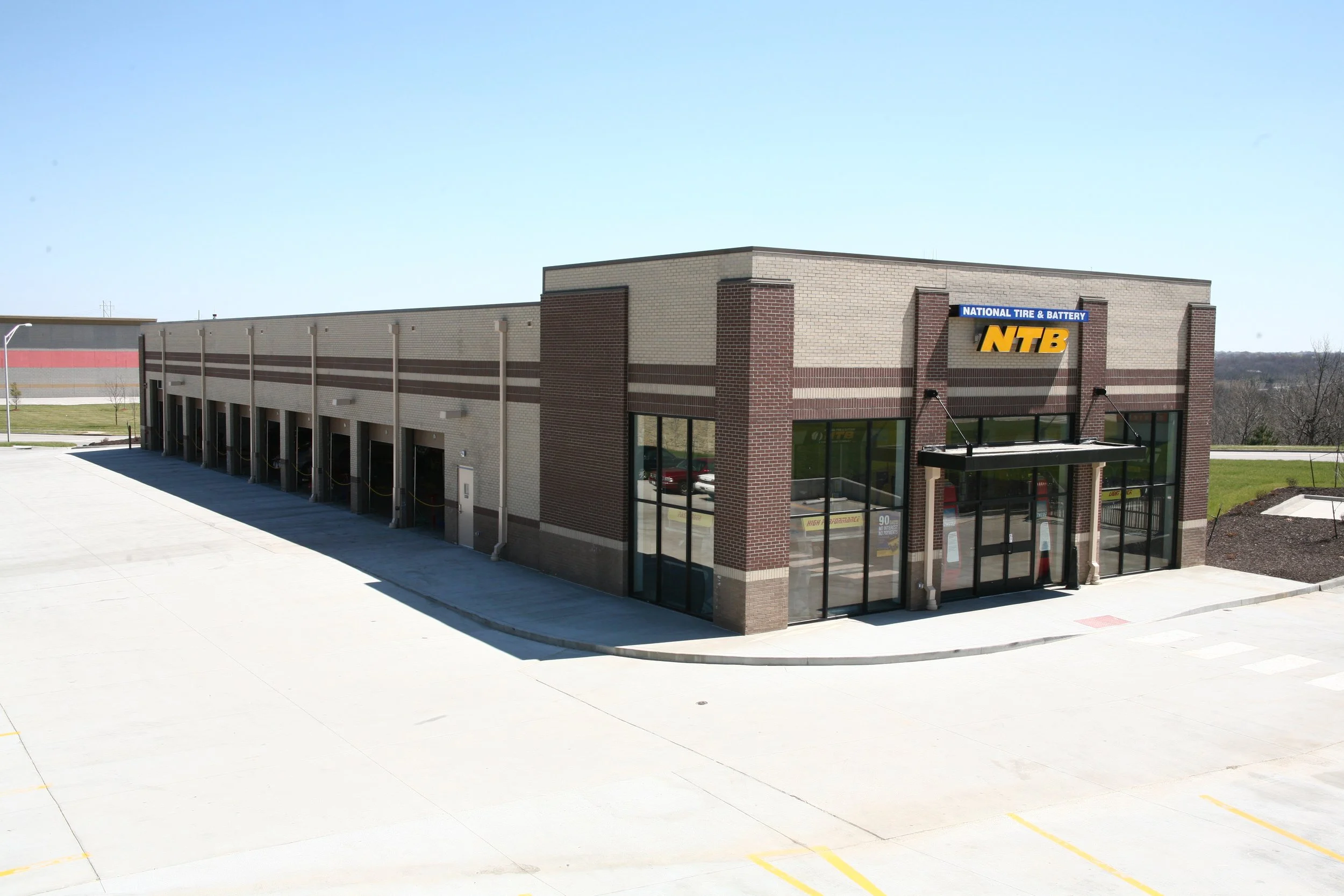 Exterior of a commercial building with a sign for National Tire & Battery (NTB), featuring large glass windows at the front and a parking lot in the foreground.