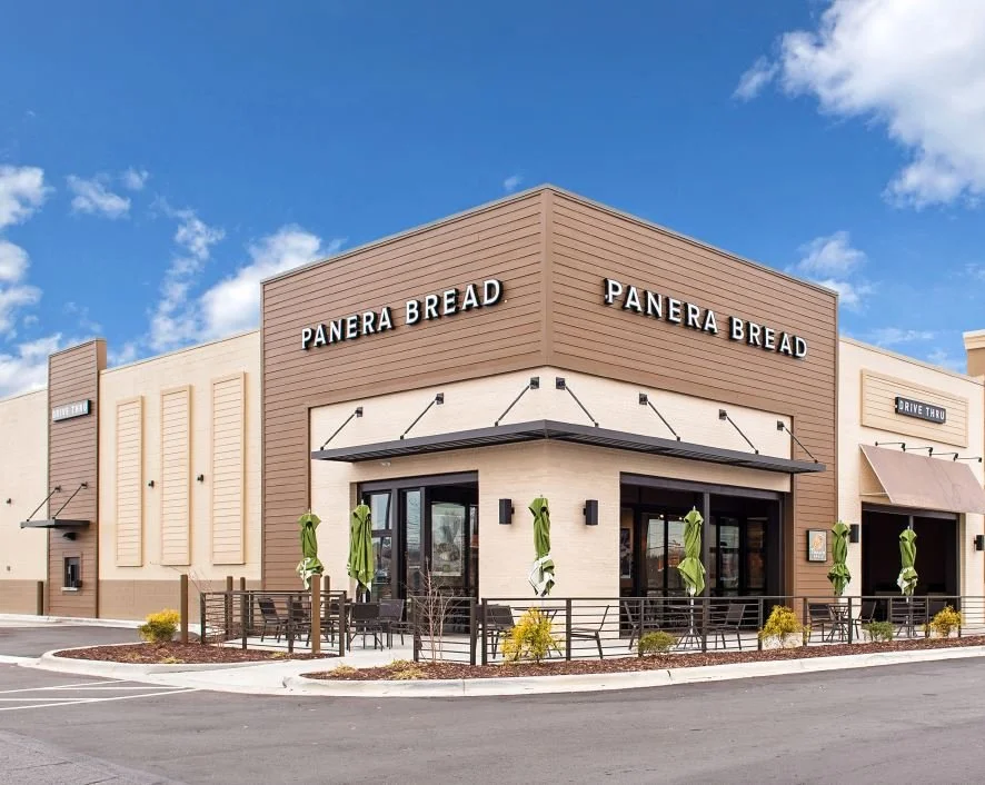Exterior of Panera Bread bakery-café with outdoor seating, umbrellas, and clear blue sky.