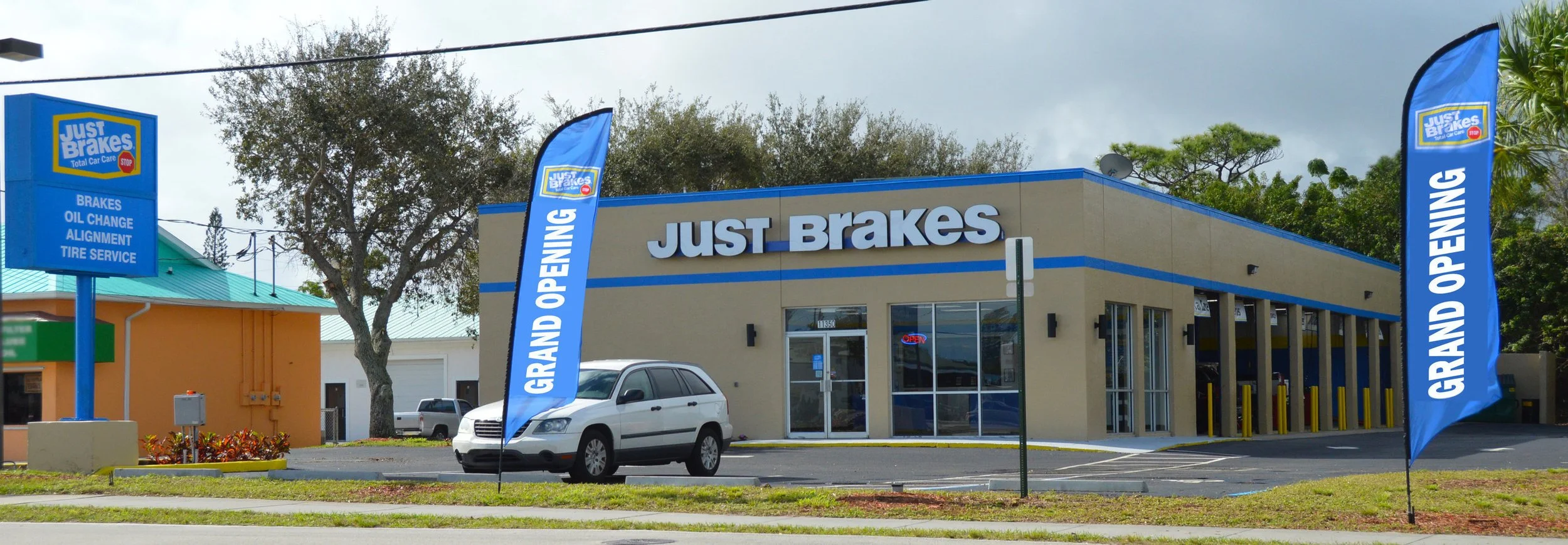 Exterior of a car service shop called Just Brakes with a sign listing services like brakes, oil change, alignment, and tire service. Two vertical blue banners in front of the shop, one saying 'Grand Opening'. A silver minivan is parked near the entra