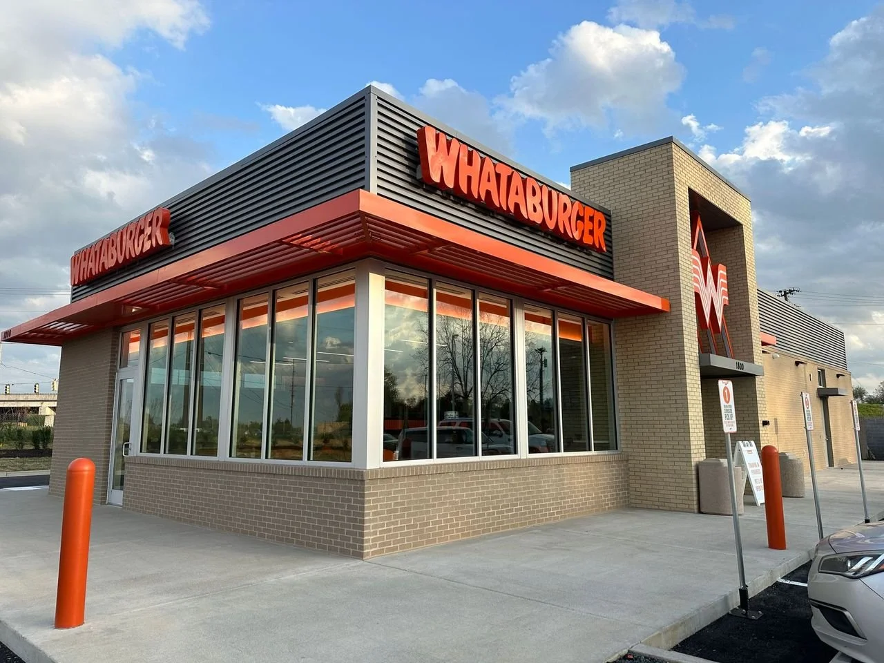 Exterior of a Whataburger fast food restaurant with large glass windows, orange signage, and a drive-thru lane, under a partly cloudy sky.