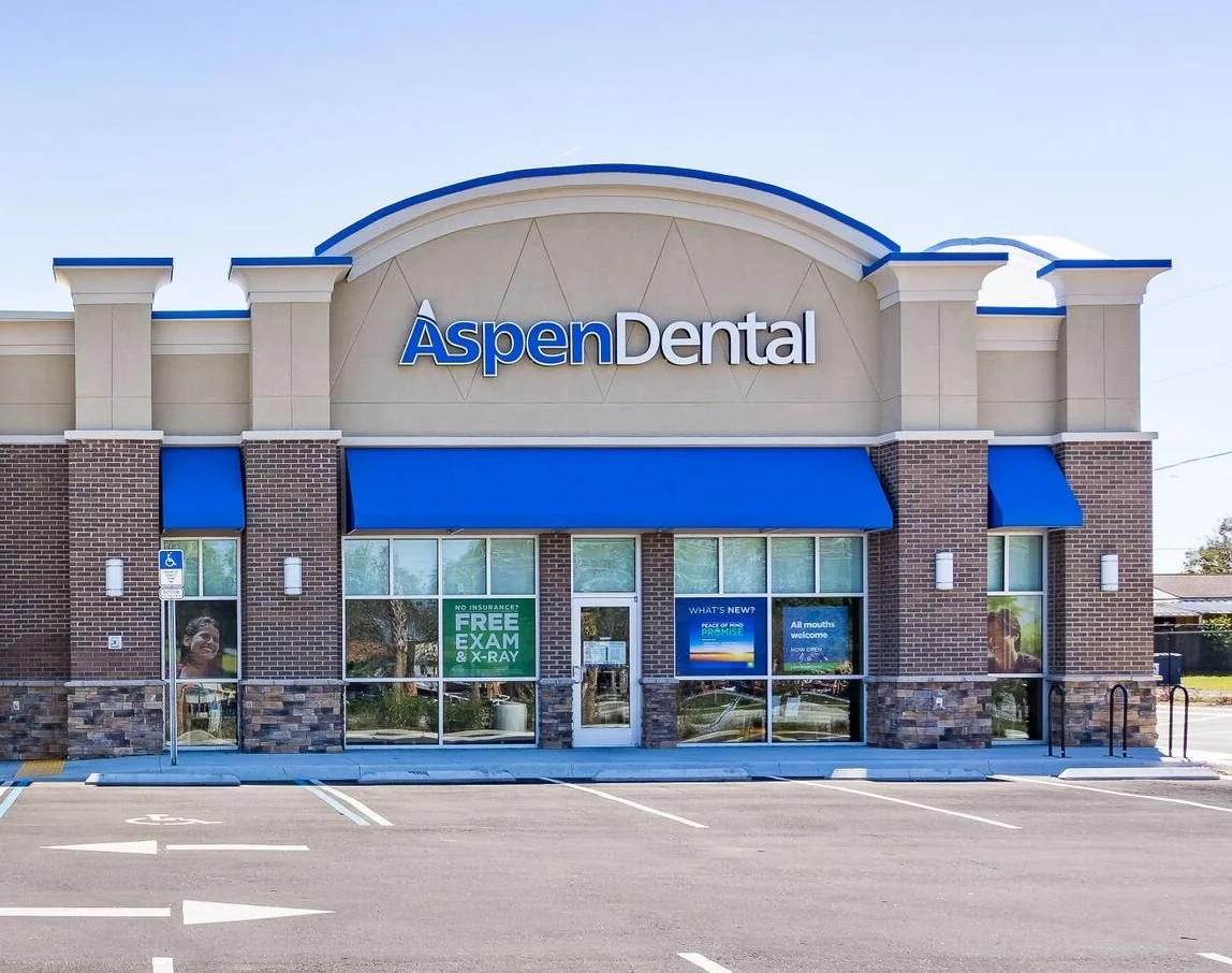Front view of Aspen Dental clinic building with blue awnings and signage, in a parking lot with designated spaces, some posters visible in windows advertising free exams and ongoing promotions.