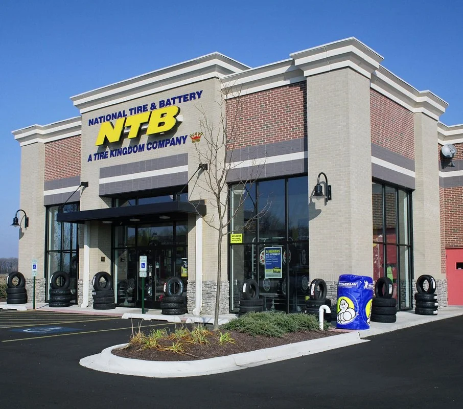 Exterior of a tire and battery store named National Tire & Battery (NTB), with tires displayed outside, a parking lot, and a leafless tree in front.